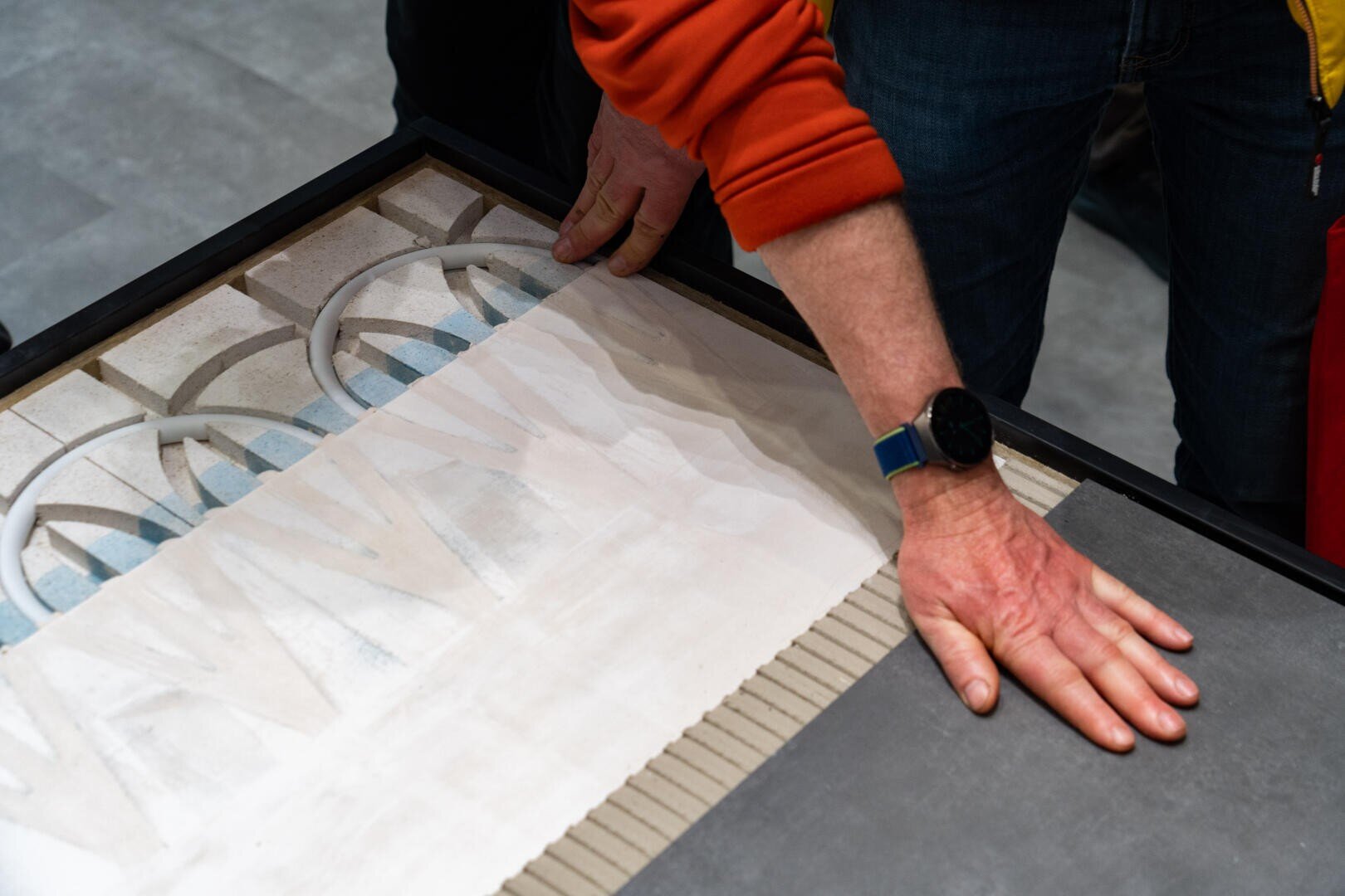 Eine Person, die eine Smartwatch und eine orangefarbene Hülle trägt, passt mit beiden Händen dekorative Kacheln auf einer Oberfläche an, indem sie komplizierte Muster neben einfarbigen Kacheln anordnet.