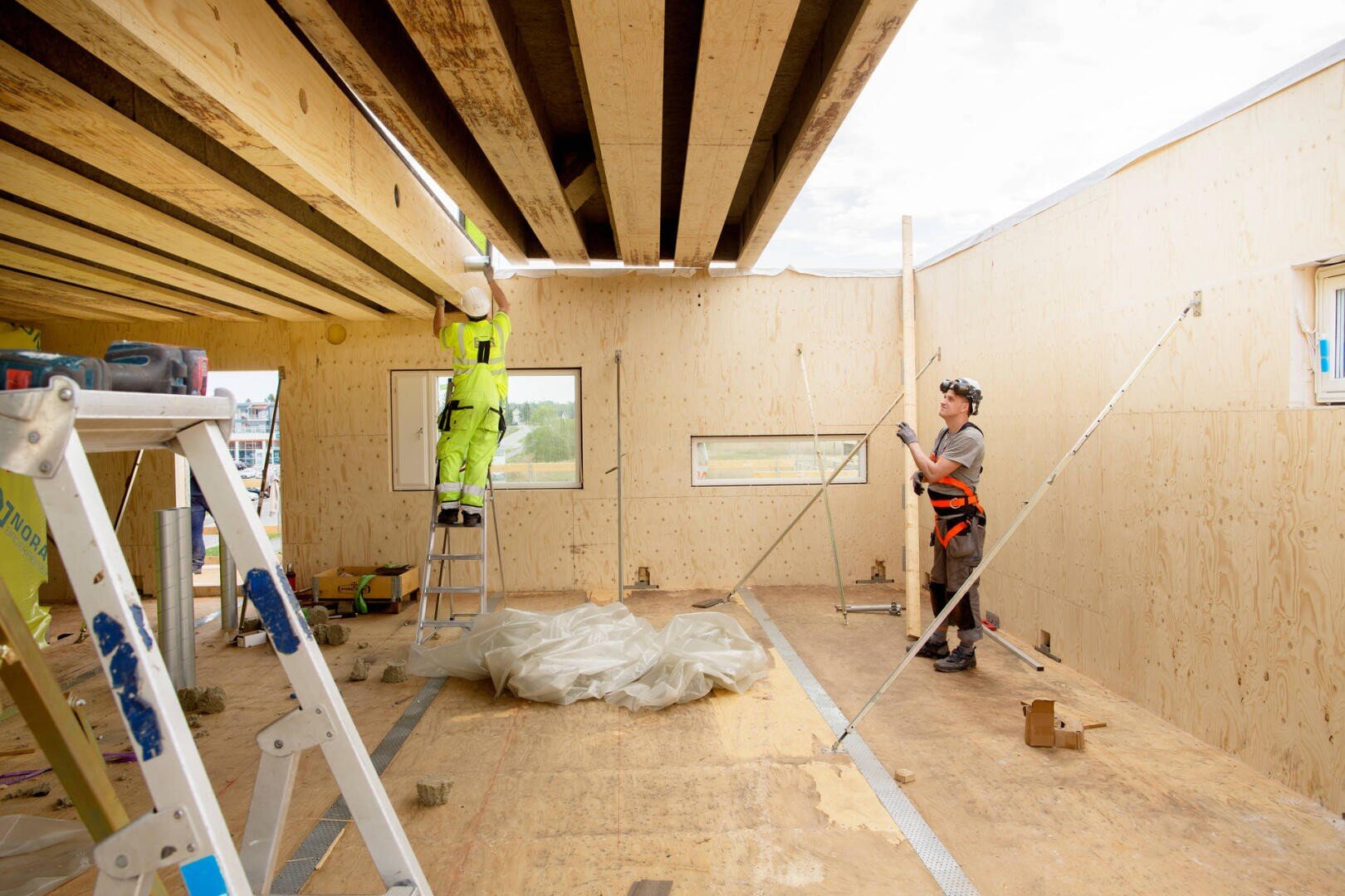 Two construction workers are building a wooden structure; one stands on a ladder and attaches beams to the ceiling, while the other stands on the floor with tools and measures or checks vertical supports. The room is unfinished and well lit.