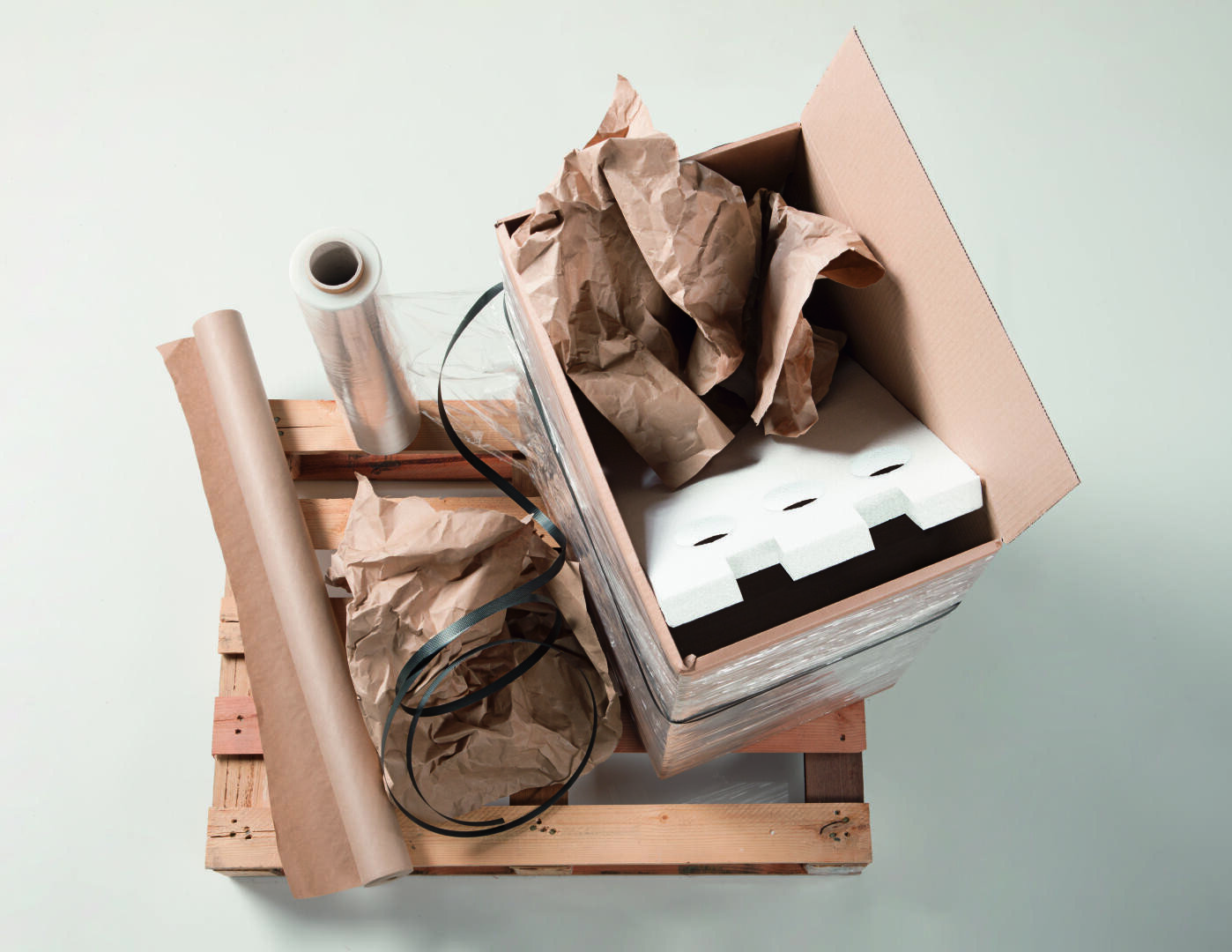 Ein mit braunem Papier, weißem Schaumstoff und schwarzen Bändern gefüllter Karton steht von oben gesehen auf einer Holzpalette neben einer Rolle Plastikfolie und einem Blatt braunem Papier.
