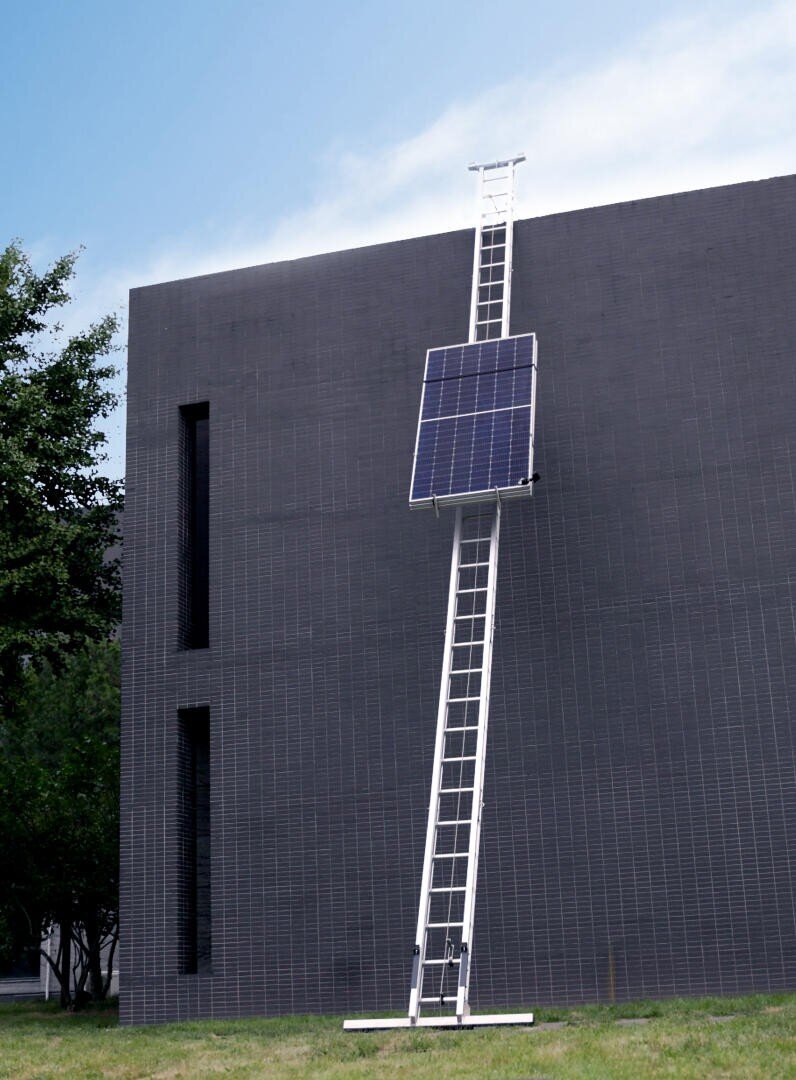 A tall white ladder is leaning against a dark gray building, with a large solar panel mounted near the top of the ladder. Green grass and a tree are visible to the left, under a partly cloudy sky.