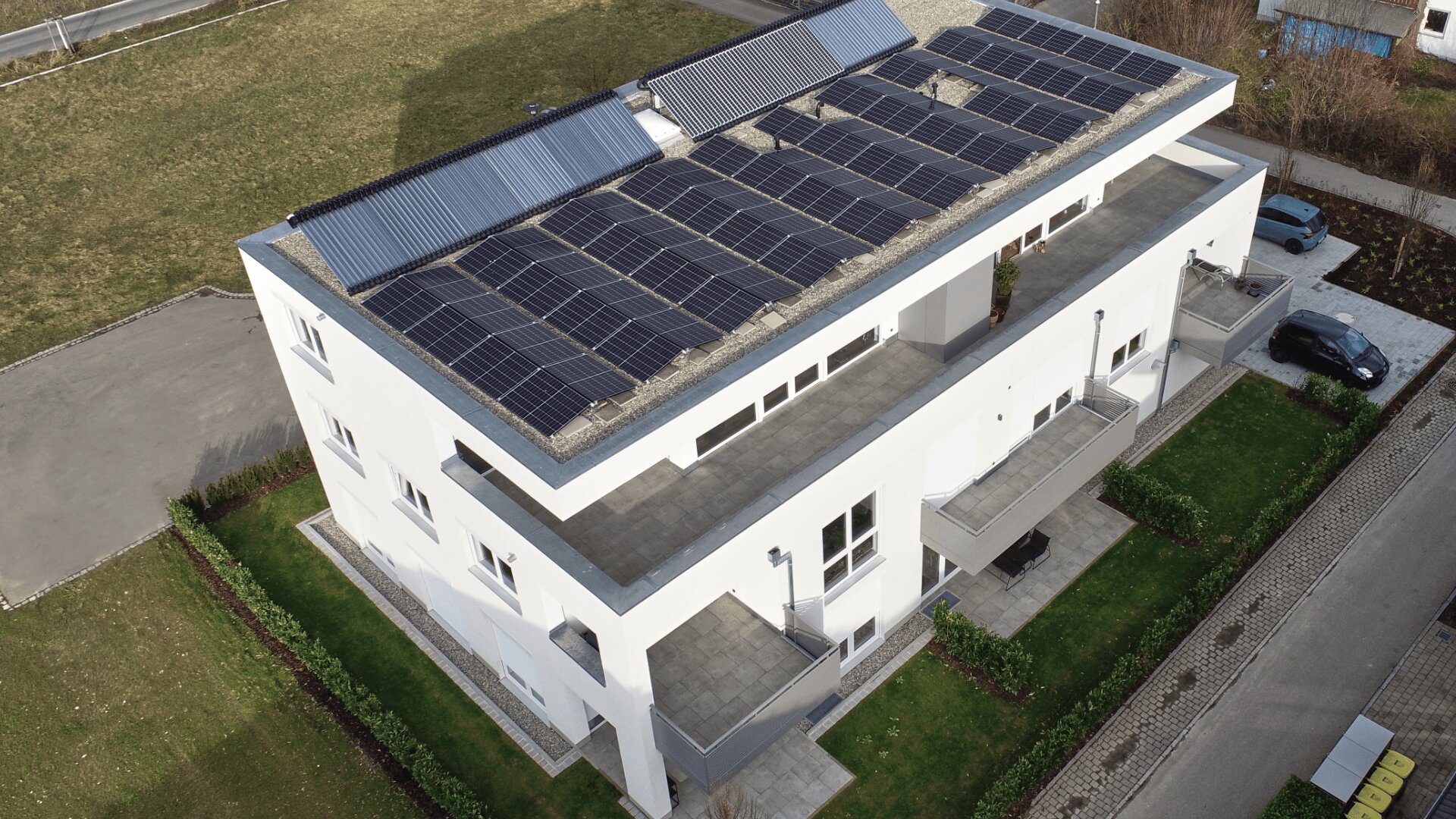 Ein modernes weißes Gebäude mit mehreren Balkonen und Solarmodulen, die das gesamte Flachdach bedecken, umgeben von grünen Rasenflächen und gepflasterten Einfahrten.