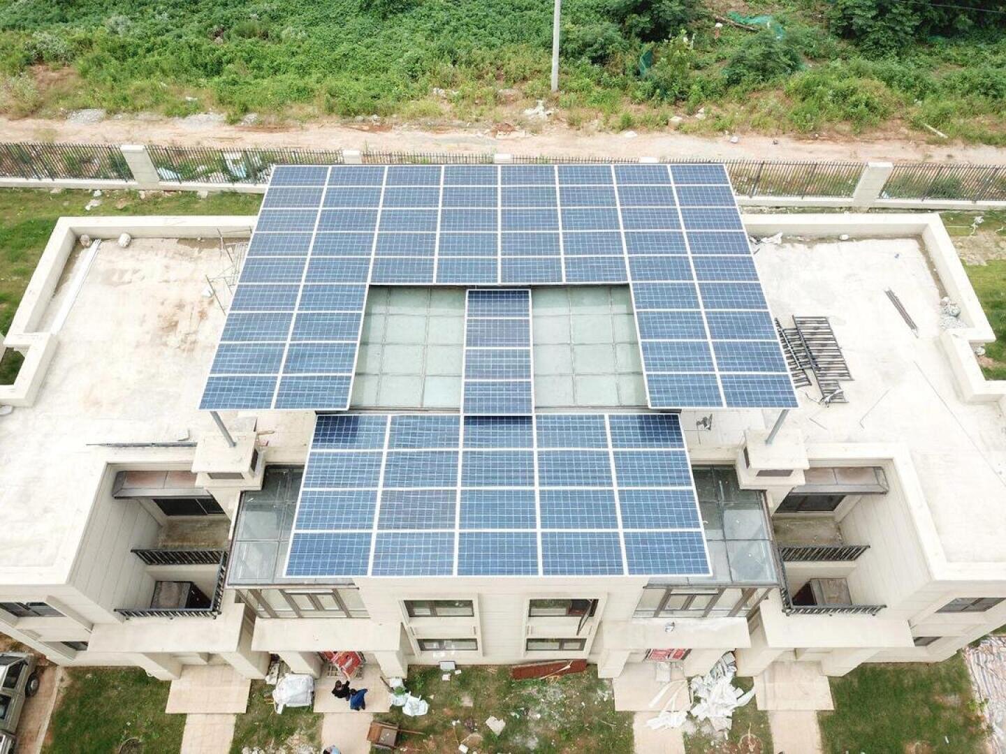 Luftaufnahme eines Gebäudes, auf dessen flachem Dach ein großes rechteckiges Netz von Solarmodulen installiert ist, umgeben von grünem Gras und einem eingezäunten Bereich mit Vegetation.