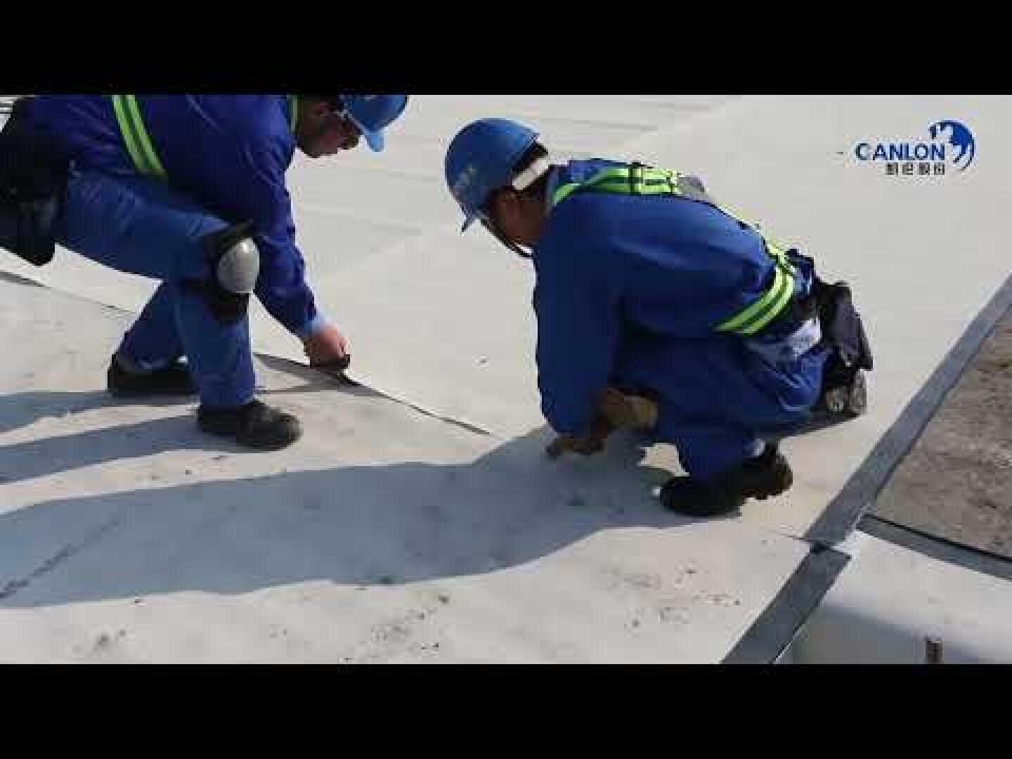 Two construction workers in blue uniforms, helmets, and safety vests are kneeling and working together on a flat rooftop, handling a large sheet of material. The logo CANLON is visible in the top right corner.