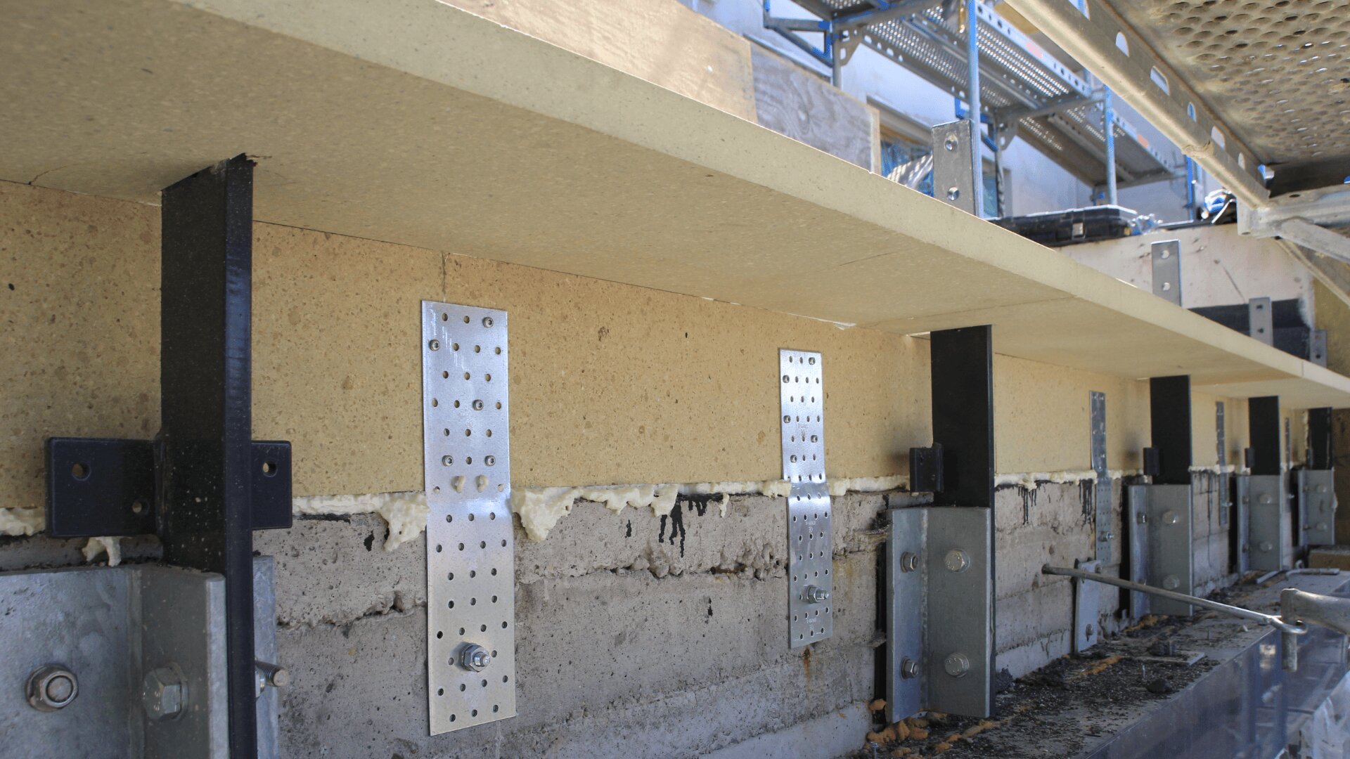 Nahaufnahme eines im Bau befindlichen Gebäudes mit an Isolierung und Beton befestigten Metallklammern und -stützen, darüber sind Gerüst und Baumaterialien sichtbar.