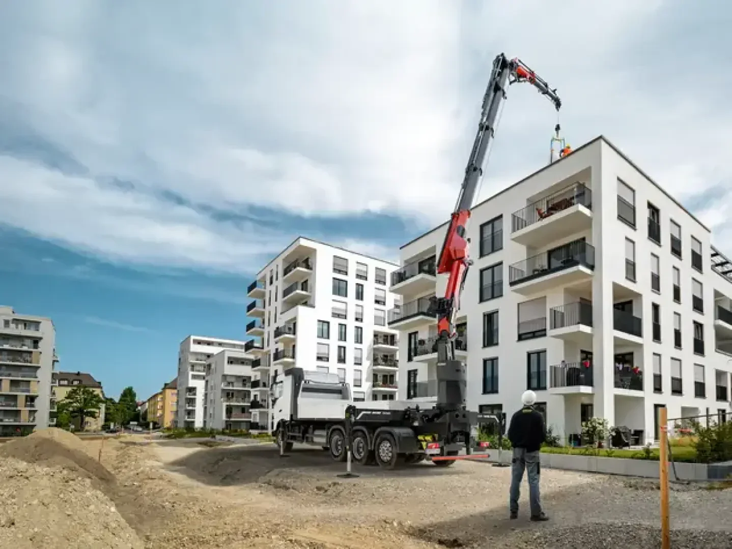Ein Bauarbeiter mit Helm beobachtet einen Lkw mit einem Kran, der neben modernen weißen Wohngebäuden bei teilweise bewölktem Himmel Materialien anhebt. Im Vordergrund sind Erdhaufen und Bauzonen zu sehen.