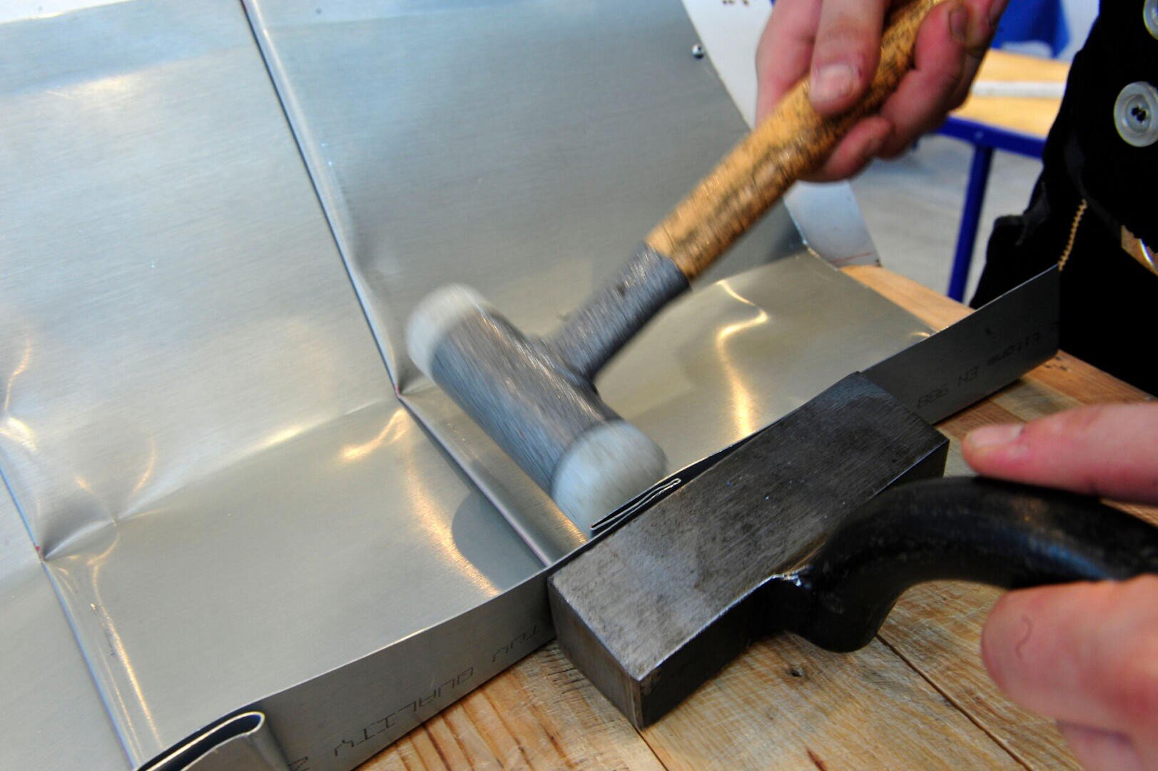 A person uses a mallet and a metal tool to shape or bend a sheet of metal on a wooden surface.