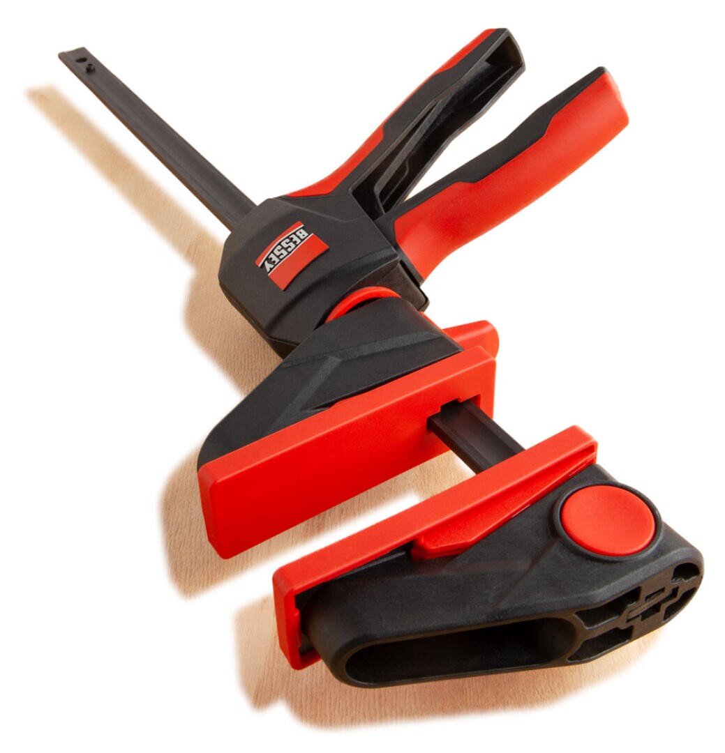 A red and black bar clamp with a trigger handle, designed for woodworking and holding materials tightly together, positioned on a light surface.