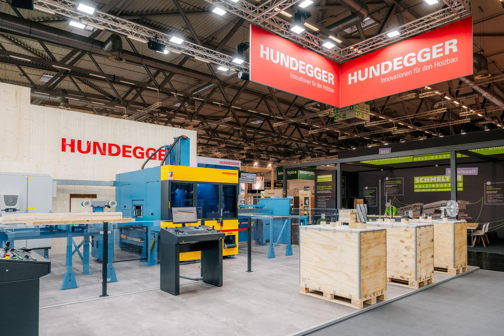 Ausstellungsstand der Firma Hundegger mit großen industriellen Holzbearbeitungsmaschinen, Ausstellungstischen mit Holzkisten und Informationstafeln in einer geräumigen, gut beleuchteten Messehalle.