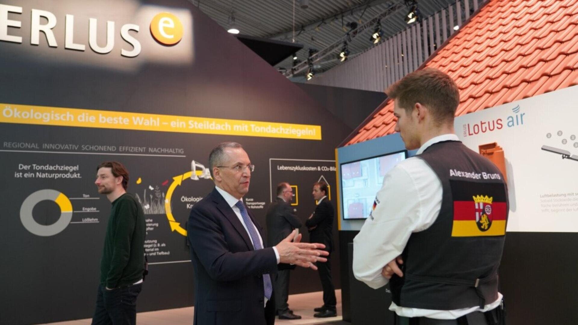 Two men talk at a trade show booth about eco-friendly roofing tiles. One man wears a suit; the other wears a uniform with a German flag patch and “Alexander Bruns.” A display with product info and a roof model is in the background.