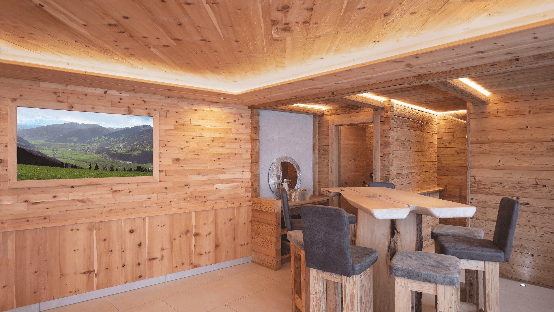 Ein gemütlicher Raum mit holzgetäfelten Wänden, Decken und Möbeln. Es gibt einen Holztisch mit gepolsterten Stühlen, einen runden Spiegel und ein gerahmtes Landschaftsfoto von Bergen und grünen Feldern an der Wand. Warmes Licht sorgt für eine einladende Atmosphäre.