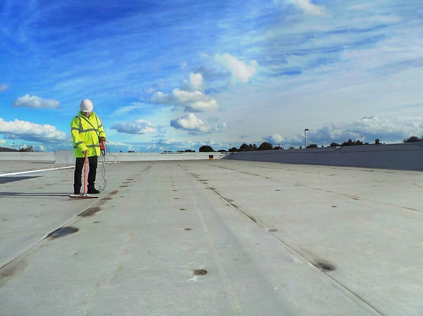 A person in a high-visibility vest and hard hat inspects a large, flat roof with a handheld device under a bright, partly cloudy sky. The roof stretches into the distance.