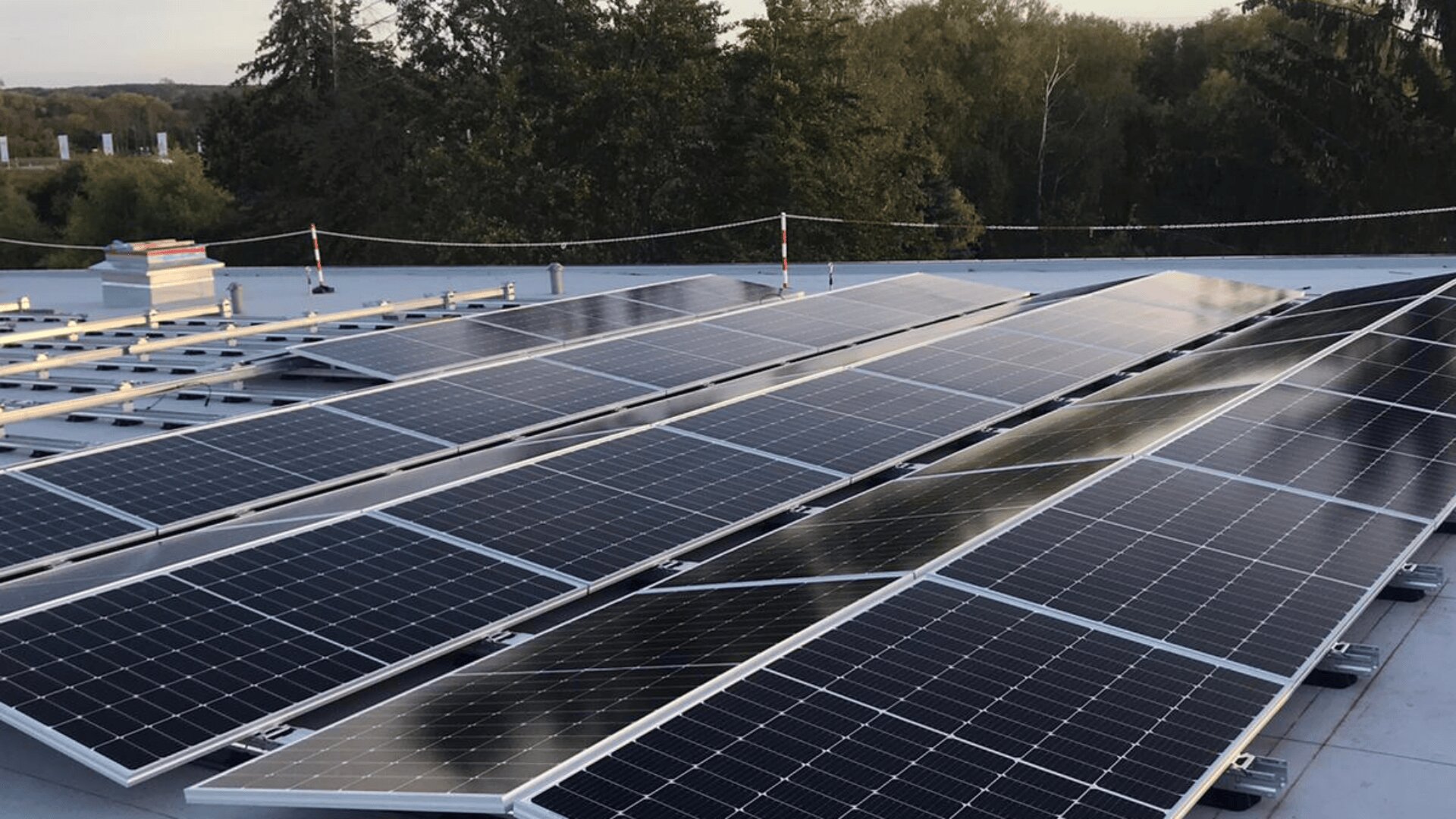 Rows of solar panels are installed on a flat rooftop, surrounded by trees and under a partly cloudy sky. The panels are angled to capture sunlight for energy generation.
