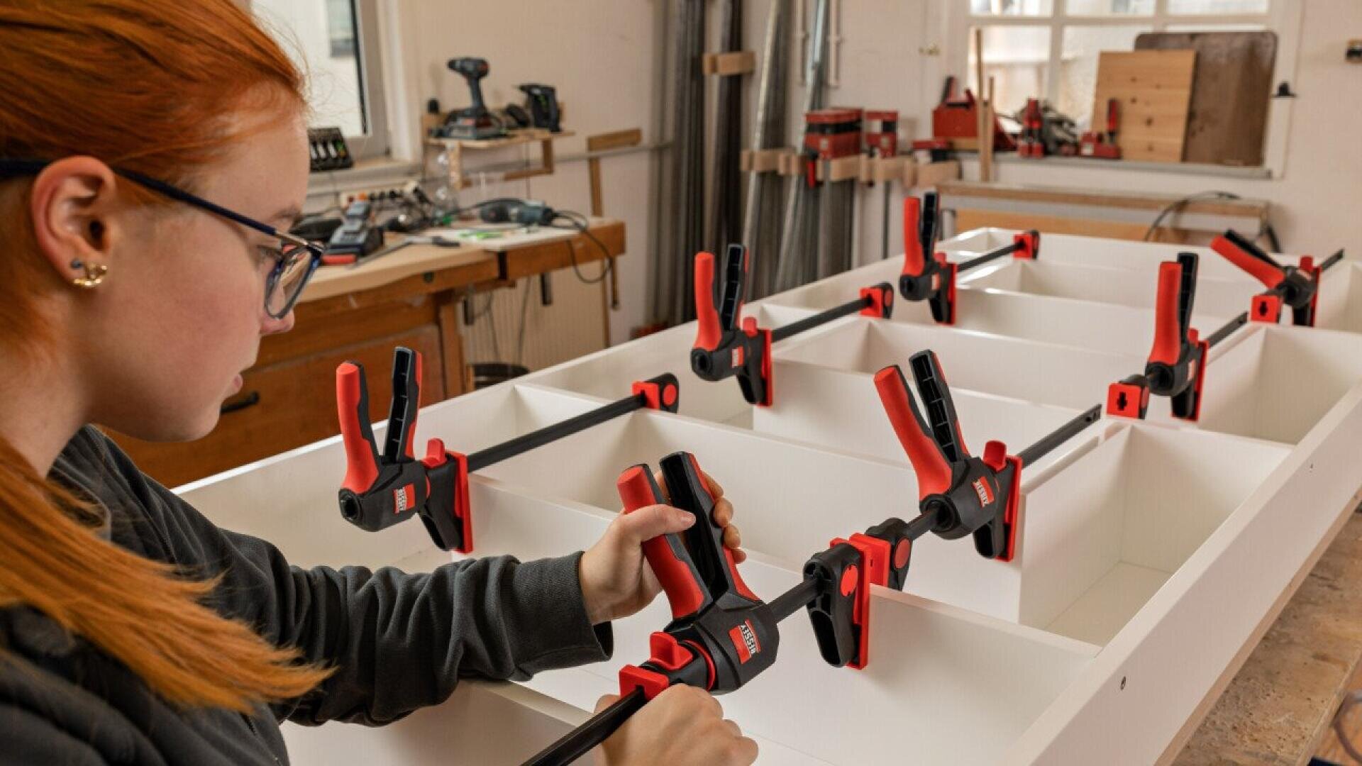 A person with red hair uses large bar clamps to secure pieces of white wood in a workshop, assembling a piece of furniture. Tools and materials are visible in the background.
