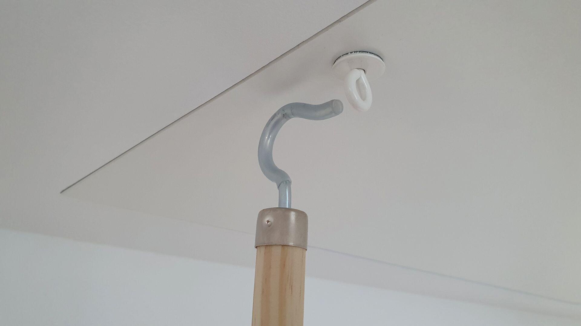 A metal hook attached to a wooden pole is being used to pull down a small handle on a ceiling hatch or attic door. The white ceiling and hatch panel are visible in the background.