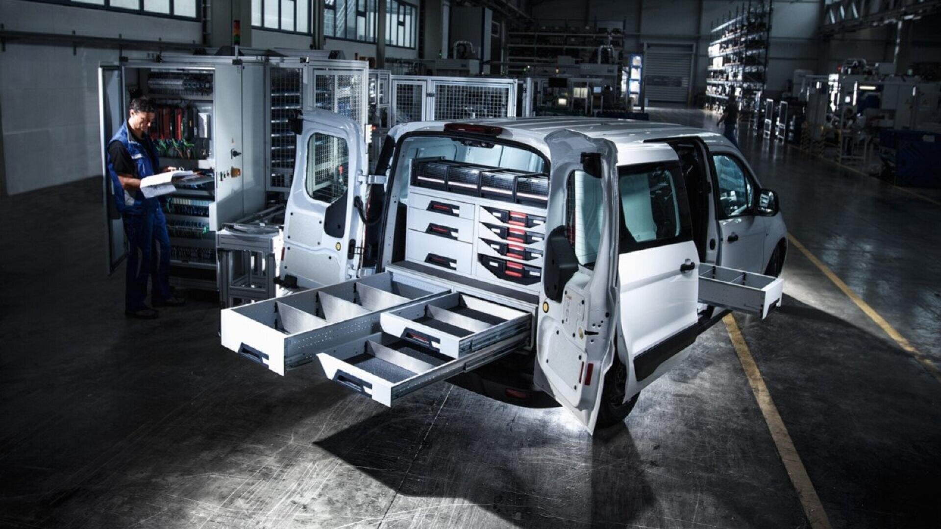 A white van with its rear and side doors open displays multiple organized storage drawers and shelves. A person stands nearby in a warehouse, inspecting items on a clipboard.