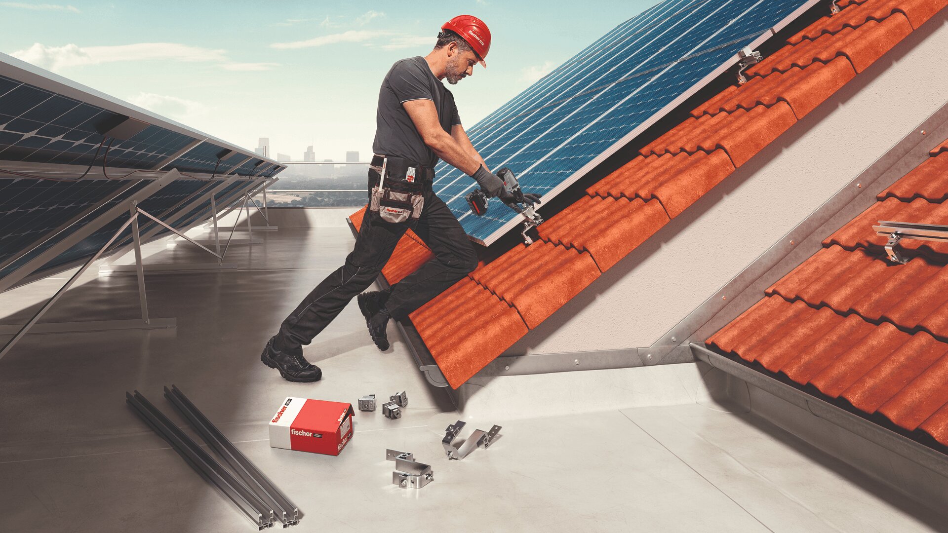 Ein Arbeiter mit einem roten Schutzhelm installiert Montagehalterungen auf einem roten Ziegeldach neben Solarmodulen. Auf dem Dach neben ihm liegen Installationswerkzeuge, Metallstangen und eine Kiste mit Ersatzteilen.