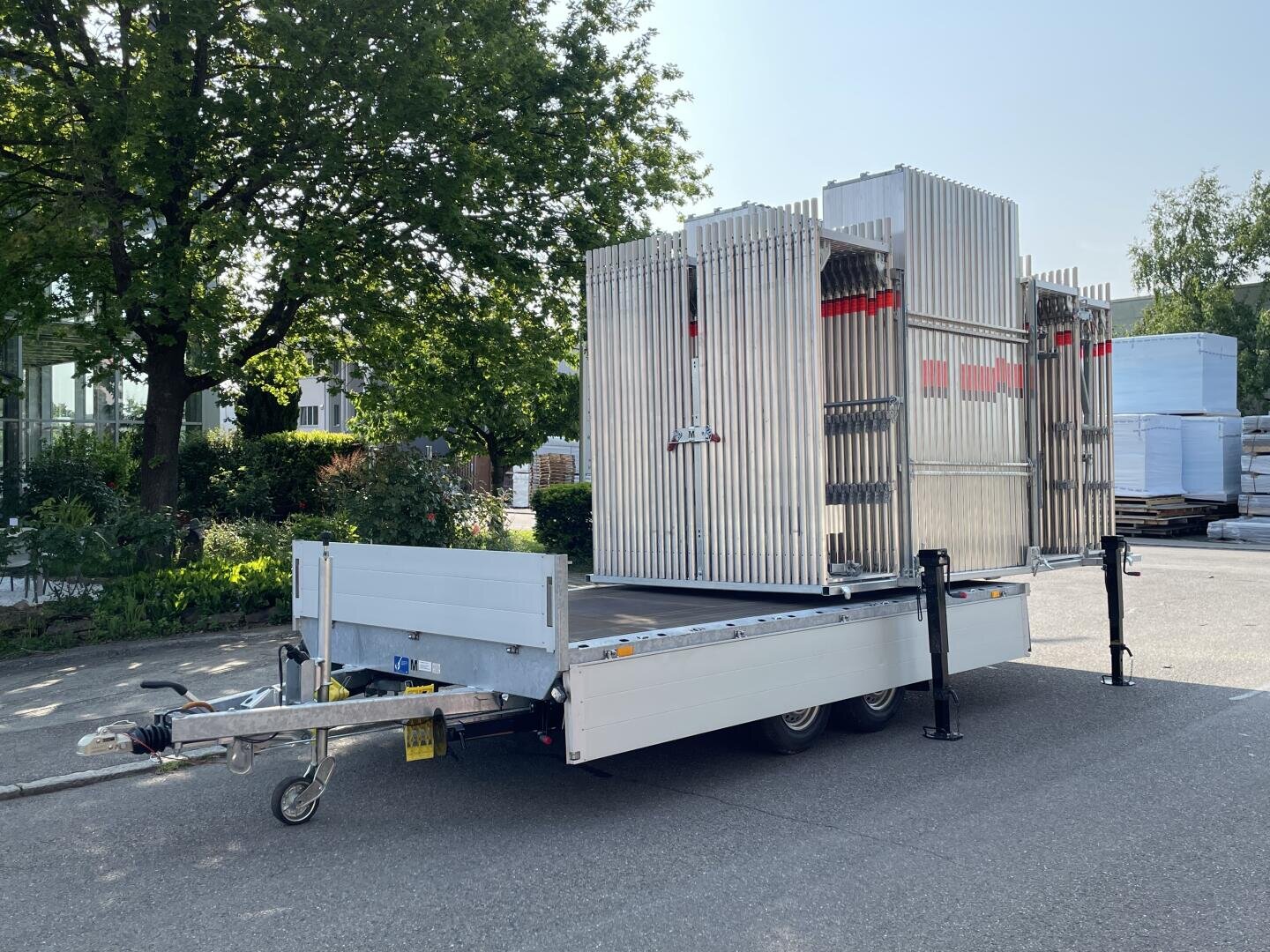 An einem sonnigen Tag steht ein mit großen Metallzaunelementen beladener Pritschenanhänger im Freien, umgeben von Bäumen und Industriegebäuden.