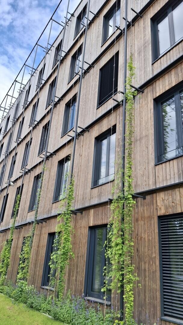A modern multi-storey building with wood paneling, large windows and vertical trellises supporting green climbing plants against a partly cloudy sky.
