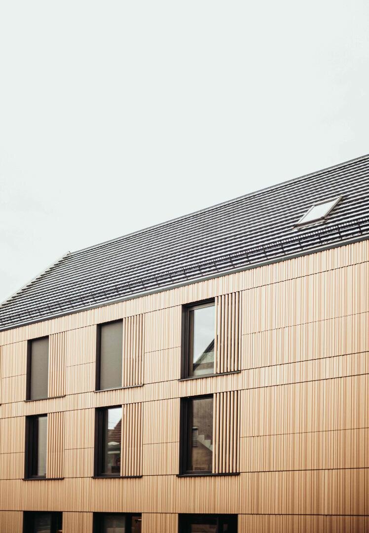 Ein modernes Gebäude mit einem schrägen, dunkelgrauen Dach und einer hellen Holzverkleidung. Große, rechteckige Fenster sind in gleichmäßigen Abständen an der Fassade unter einem bedeckten Himmel angebracht.