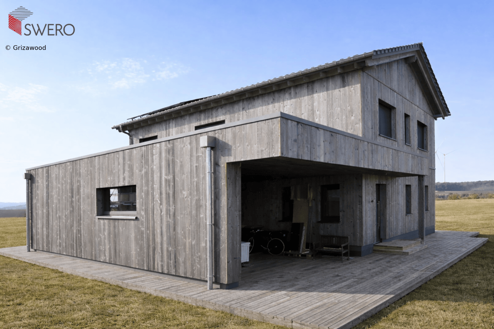 Modern two-story house made of gray wood with a flat roof extension and a covered veranda, situated on an open meadow under a partly cloudy sky.