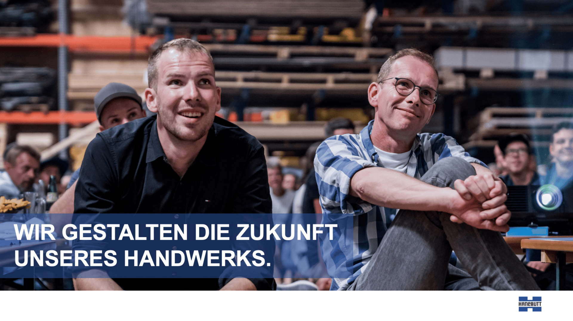 Zwei Männer sitzen lächelnd und aufmerksam in einer Werkstatt, umgeben von anderen Menschen. Der Text lautet: „Wir gestalten die Zukunft unseres Handwerks“. Im Hintergrund sind Regale und Werkzeuge zu sehen.