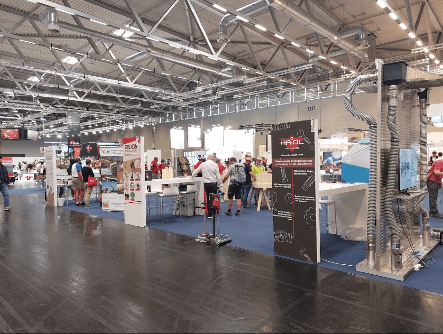 Ausstellungshalle mit verschiedenen Ständen, Bannern und Displays. Die Menschen gehen umher und tauschen sich an den Ständen aus. Der Raum hat hohe Decken mit Metallbalken, und der Boden ist teilweise mit blauen und schwarzen Abschnitten bedeckt.