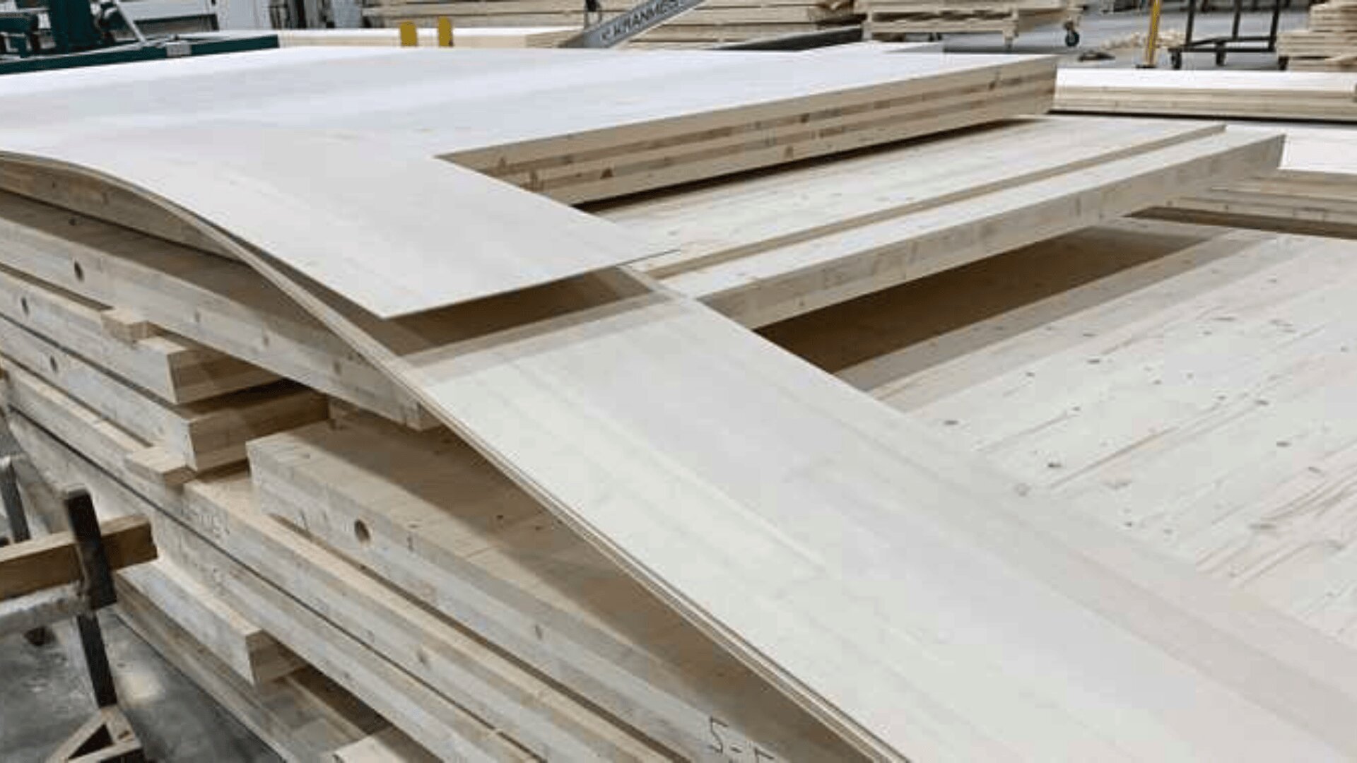 In einer Werkstatt stapeln sich helle, glatte Holzbretter und -platten. Ein flexibles Blech ist über die Oberseite des Stapels gebogen und zeigt so seine Dünnheit im Vergleich zu den dickeren Platten darunter.