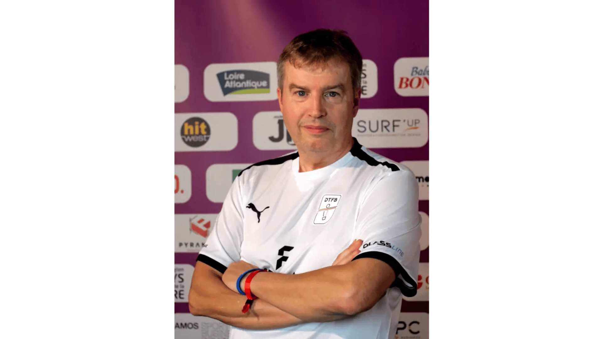 Ein Mann in einem weißen Sporttrikot mit schwarzen Akzenten steht mit verschränkten Armen vor einem violetten Hintergrund mit verschiedenen Sponsorenlogos.