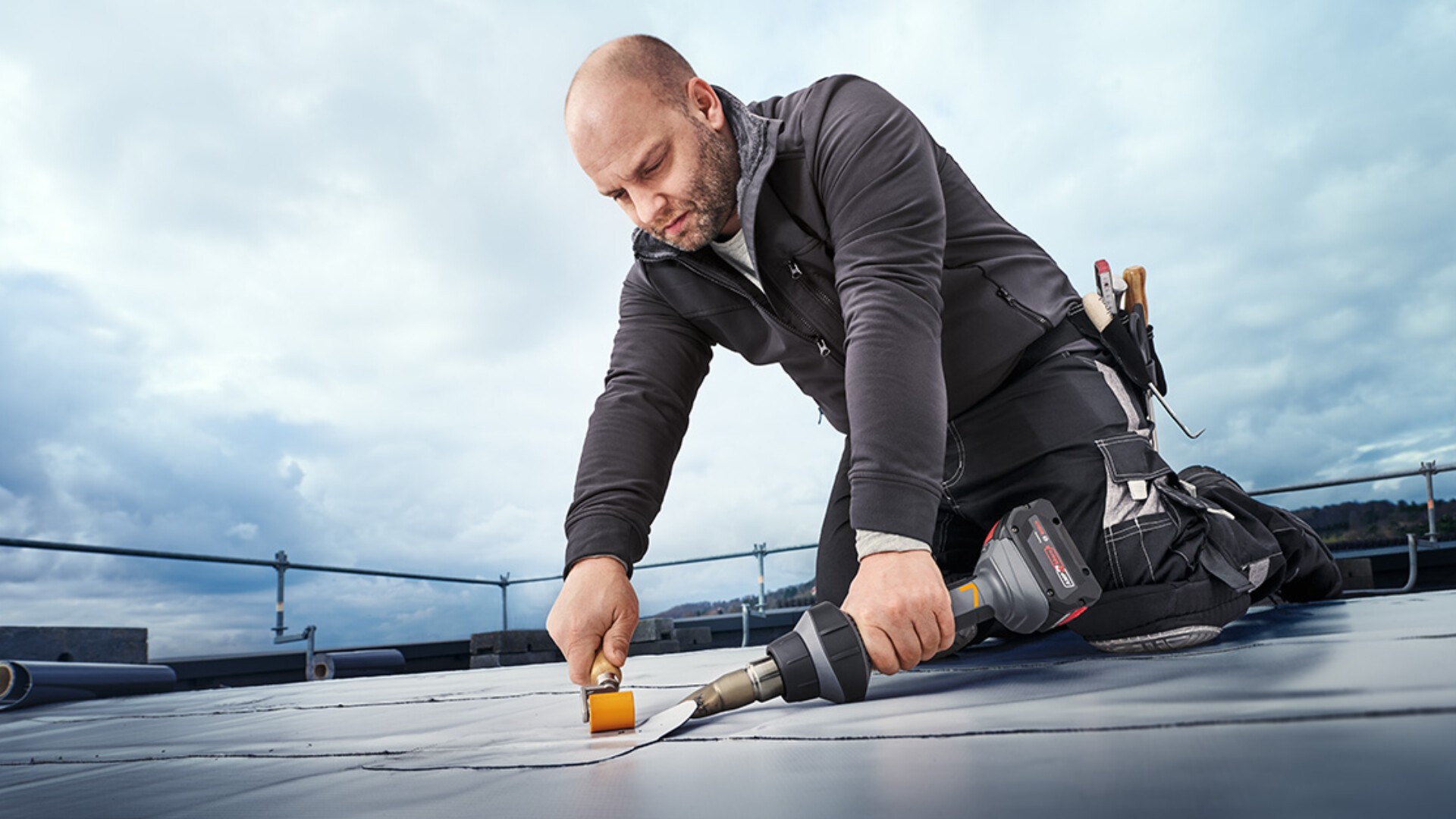 Ein Mann kniet auf einem Dach und versiegelt mit einer Heißluftpistole in der einen und einer Walze in der anderen Hand eine Naht in Dachmaterial. Er trägt Arbeitskleidung und hat Werkzeug am Gürtel. Der Himmel ist bewölkt.