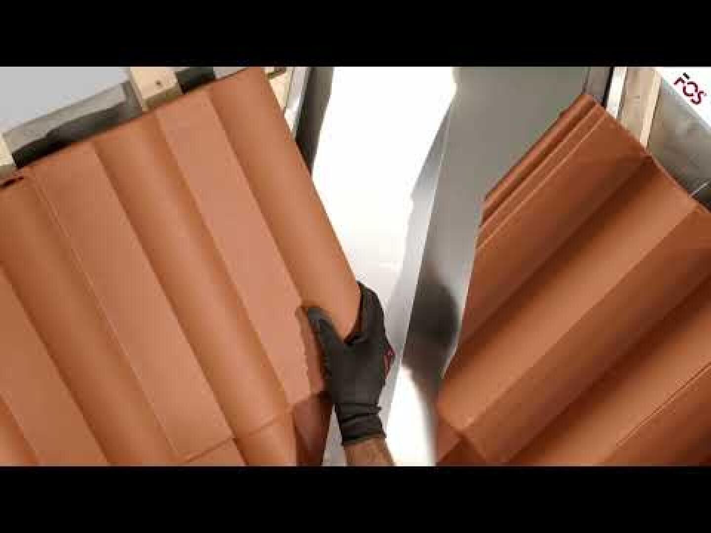 Eine Person mit einem schwarzen Handschuh hält ein braunes Blech mit einem Wellenmuster in der Nähe einer Maschine in einer Werkstatt.