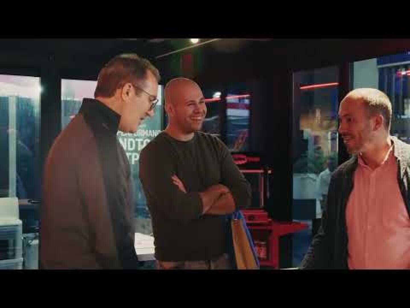 Three men are standing in a house, smiling and talking. One man looks into the camera with his arms crossed, while the other two stand at the sides and talk. The surroundings look modern with large windows in the background.