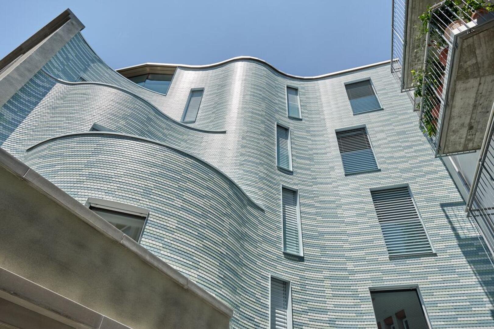 A modern building with an undulating façade covered in light blue tiles with several rectangular windows, seen from below against a clear sky. A balcony with plants can be seen on the right-hand side.