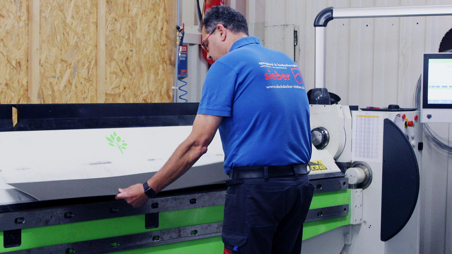 A man in a blue polo shirt operates a large industrial bending machine, carefully positioning a metal sheet. A monitor and control panel are visible on the right side of the image.