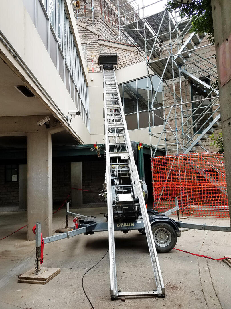 Eine Baustelle mit einem langen, geneigten mechanischen Aufzug, der vom Erdgeschoss in ein höheres Stockwerk führt. Gerüste und Sicherheitsbarrieren umgeben den Bereich, und ein Teil eines Gebäudes mit Glasfenstern ist sichtbar.
