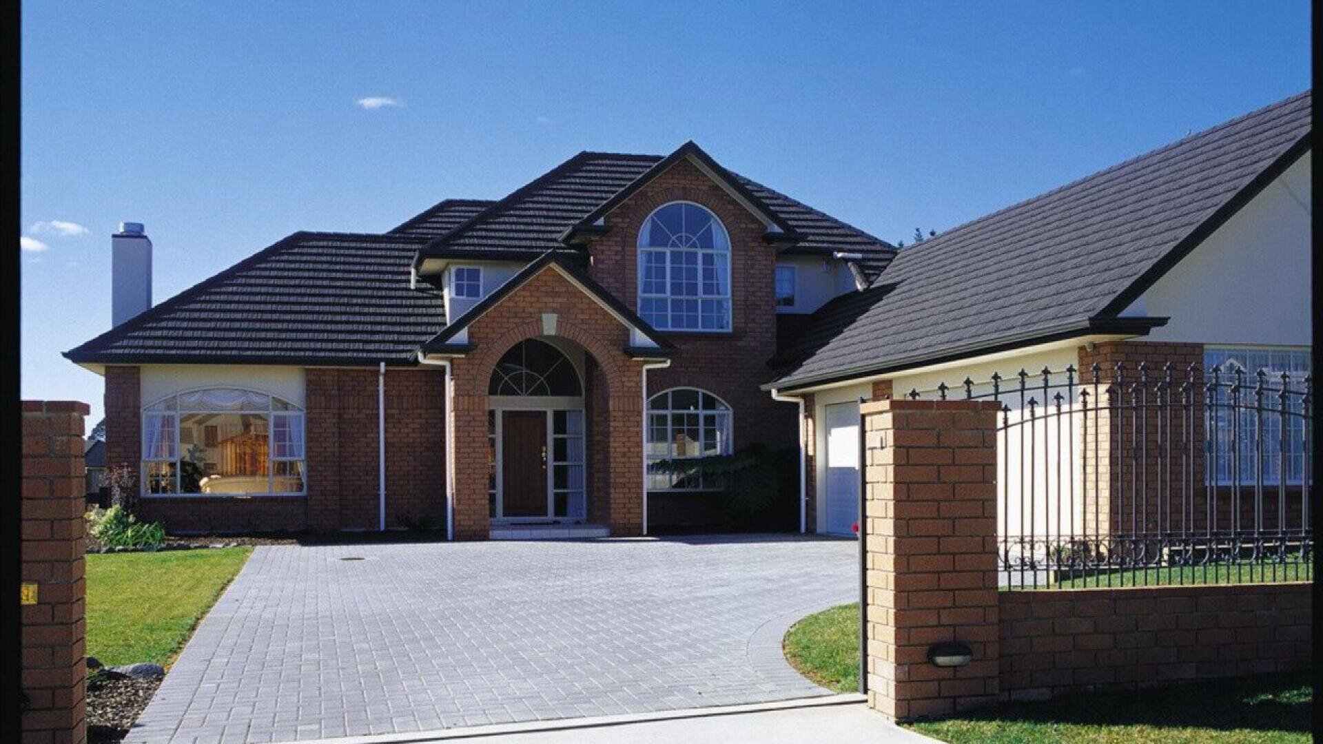 A two-story brick house with large windows, a dark tiled roof, and an attached garage, set behind a brick and wrought-iron gate on a bright, sunny day.