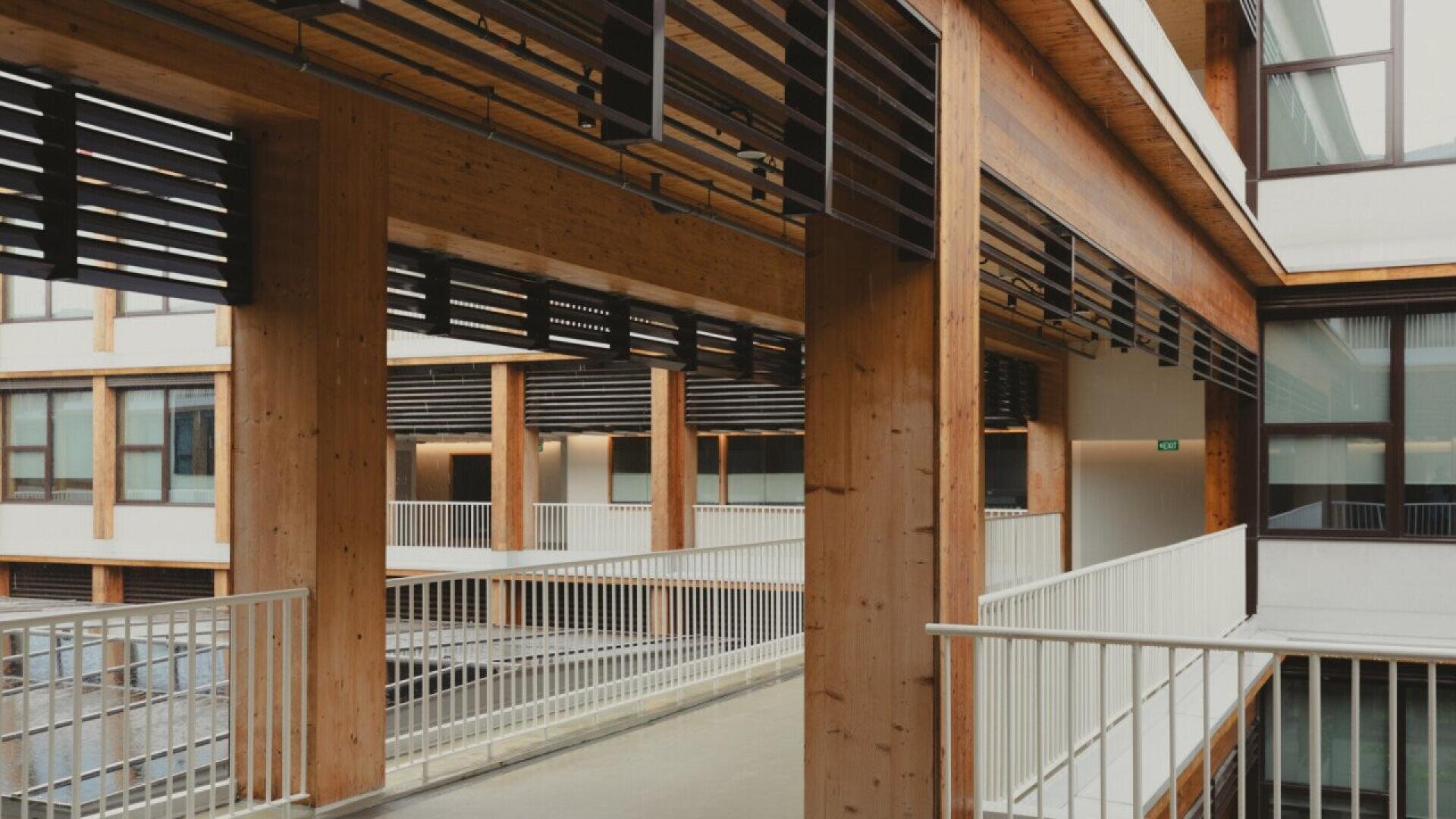 Modern building interior with exposed wooden beams, white railings, and large windows. Walkways connect different sections, creating an open and airy architectural design.