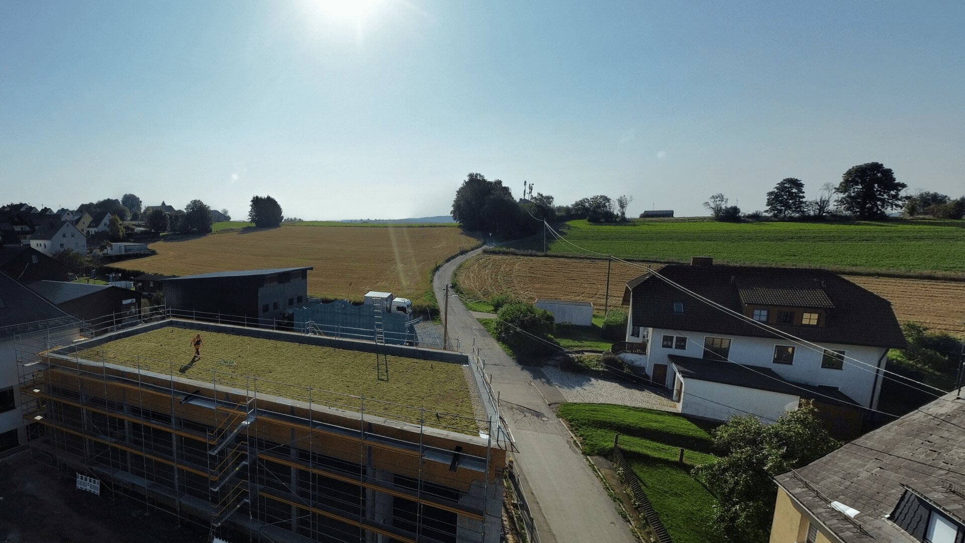 Eine ländliche Landschaft mit einer Straße, die Felder und Häuser trennt; eine Person steht auf dem grasbedeckten Dach eines im Bau befindlichen modernen Gebäudes, von oben links scheint das Sonnenlicht hinein.