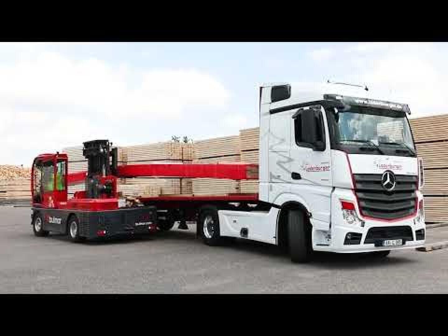 Ein weißer Mercedes-Benz-Sattelzug mit Pritschenauflieger parkt neben einem roten Gabelstapler, der einen langen Stapel Holzbretter trägt. Im Hintergrund sind unter blauem Himmel Holzbretter gestapelt.