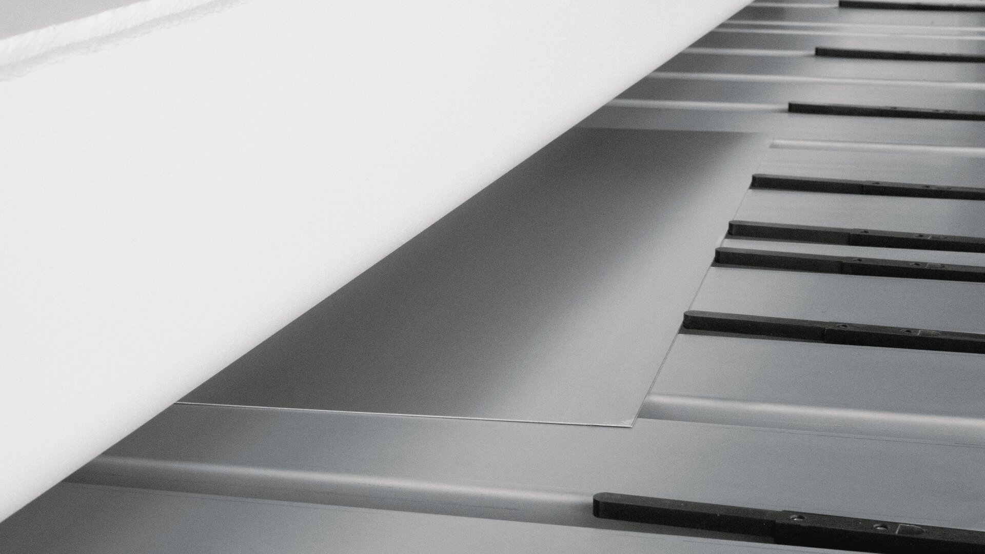 Nahaufnahme eines Metallblechs, das teilweise von einer großen Rolle weißen Materials bedeckt ist, vermutlich auf einem Förderband oder einer industriellen Verarbeitungsmaschine. Unter dem Blech sind schwarze Stützen oder Führungen sichtbar.