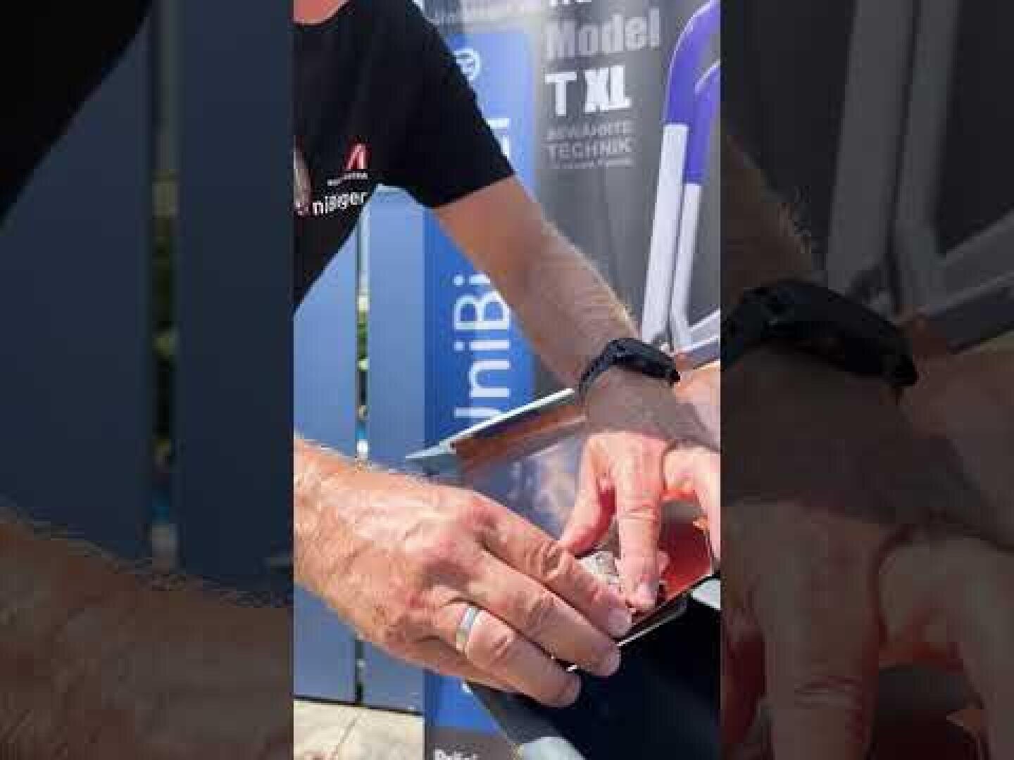 A person's hands are working on a small object outdoors next to a blue board with partially visible text. The person is wearing a black shirt and a watch.