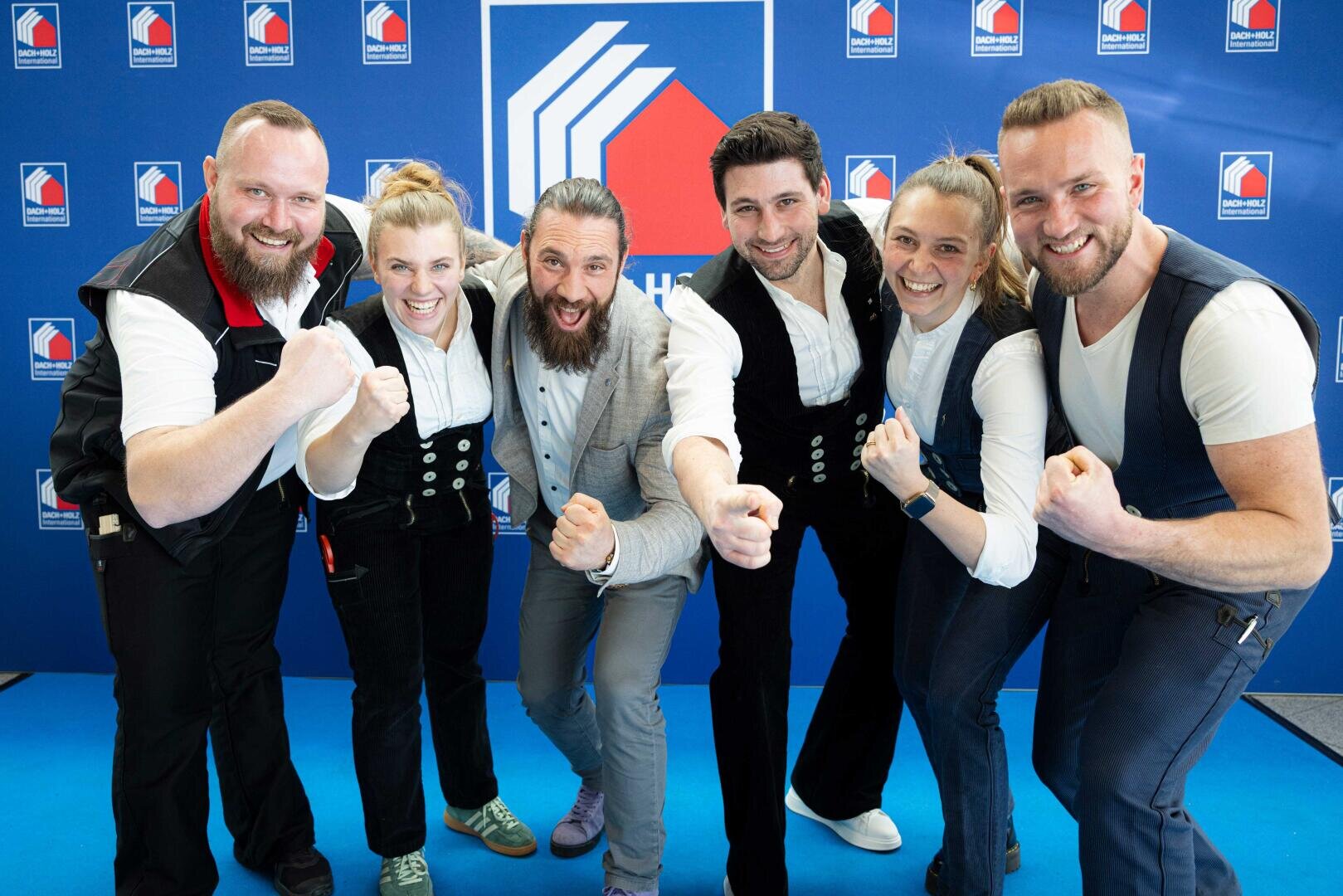 Sechs begeisterte Menschen posieren gemeinsam vor einem blauen Hintergrund mit einem hausförmigen Logo, lächeln und machen energische Gesten mit nach vorne gestreckten Fäusten.