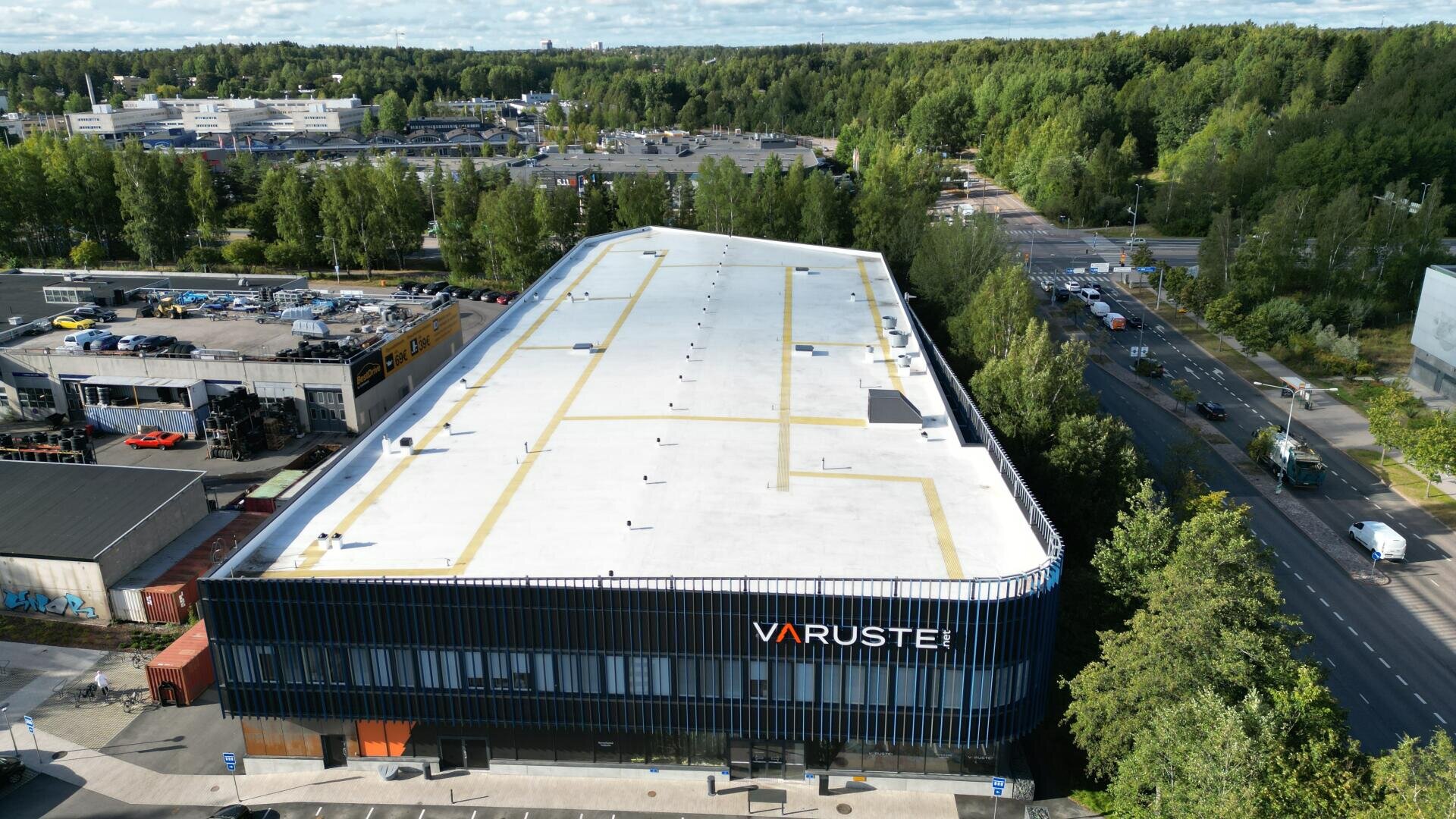 Ein großes, modernes Industriegebäude mit Varuste-Schriftzug an der Fassade, umgeben von Bäumen, Straßen und kleineren Gebäuden in der Nähe, aus der Vogelperspektive an einem teilweise bewölkten Tag.