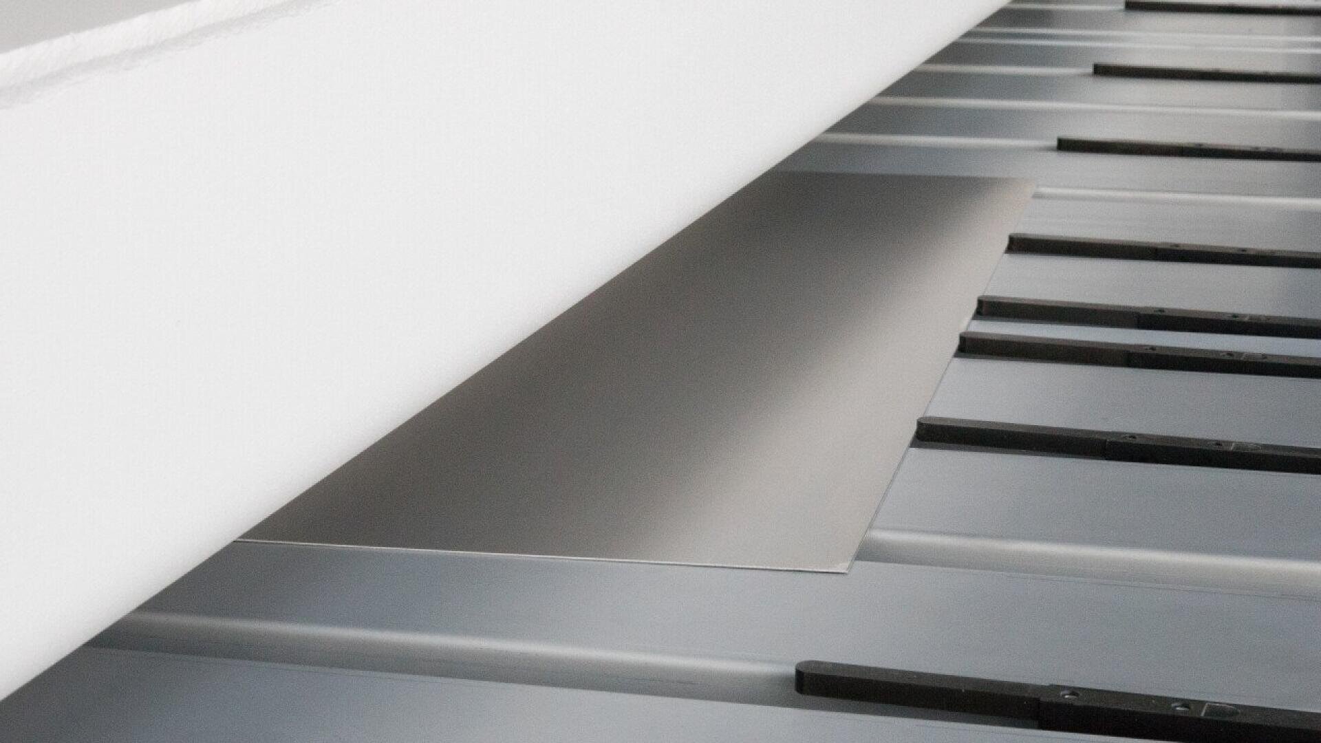 A sheet of metal is being fed into an industrial cutting or processing machine with evenly spaced black guides on a metallic surface.