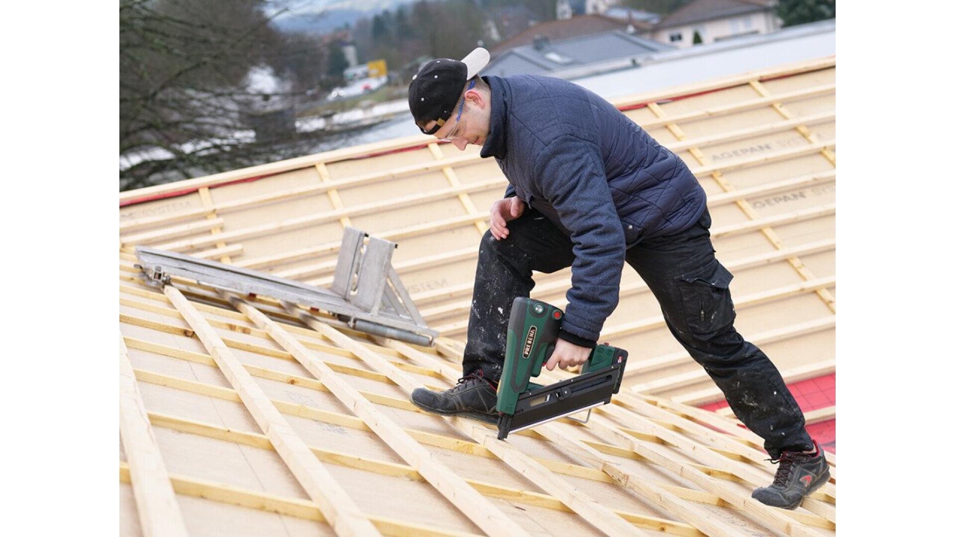 A man in work clothes and a cap uses a power tool to secure wooden beams on a sloped roof under construction. A ladder rests on the roof beside him, and houses are visible in the background.
