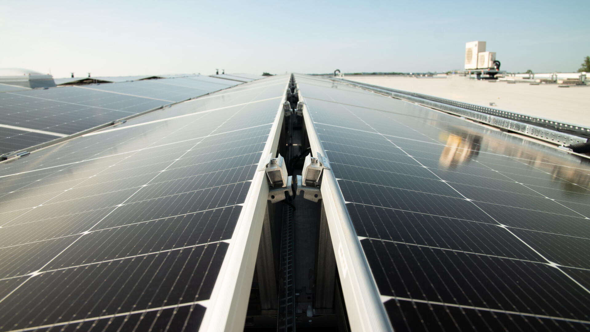 Auf einem flachen Dach erstrecken sich Reihen von Solarmodulen in die Ferne und reflektieren das Sonnenlicht unter einem klaren blauen Himmel. Die Paneele sind in geordneten Linien angeordnet und unterstreichen den Fokus auf erneuerbare Energie.