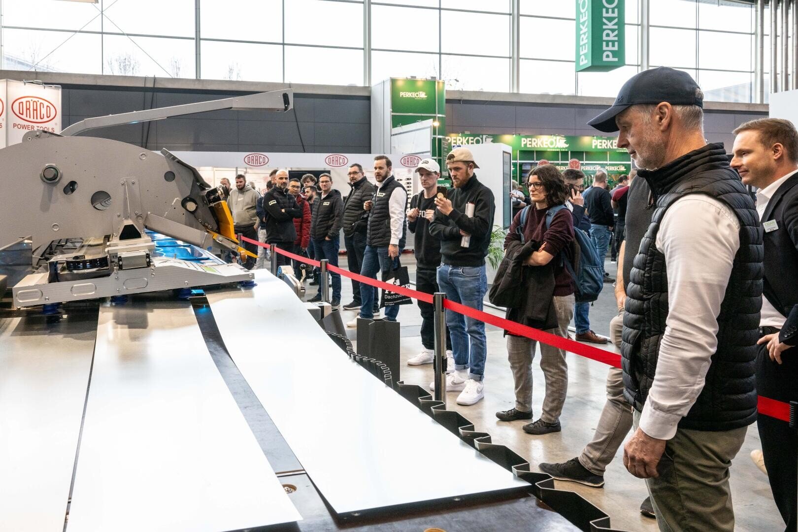 Eine Gruppe von Personen steht hinter einem roten Seil und beobachtet eine große Industriemaschine, die auf einer Ausstellung oder Messe in Betrieb ist. Einige Teilnehmer machen Fotos, während andere aufmerksam beobachten.