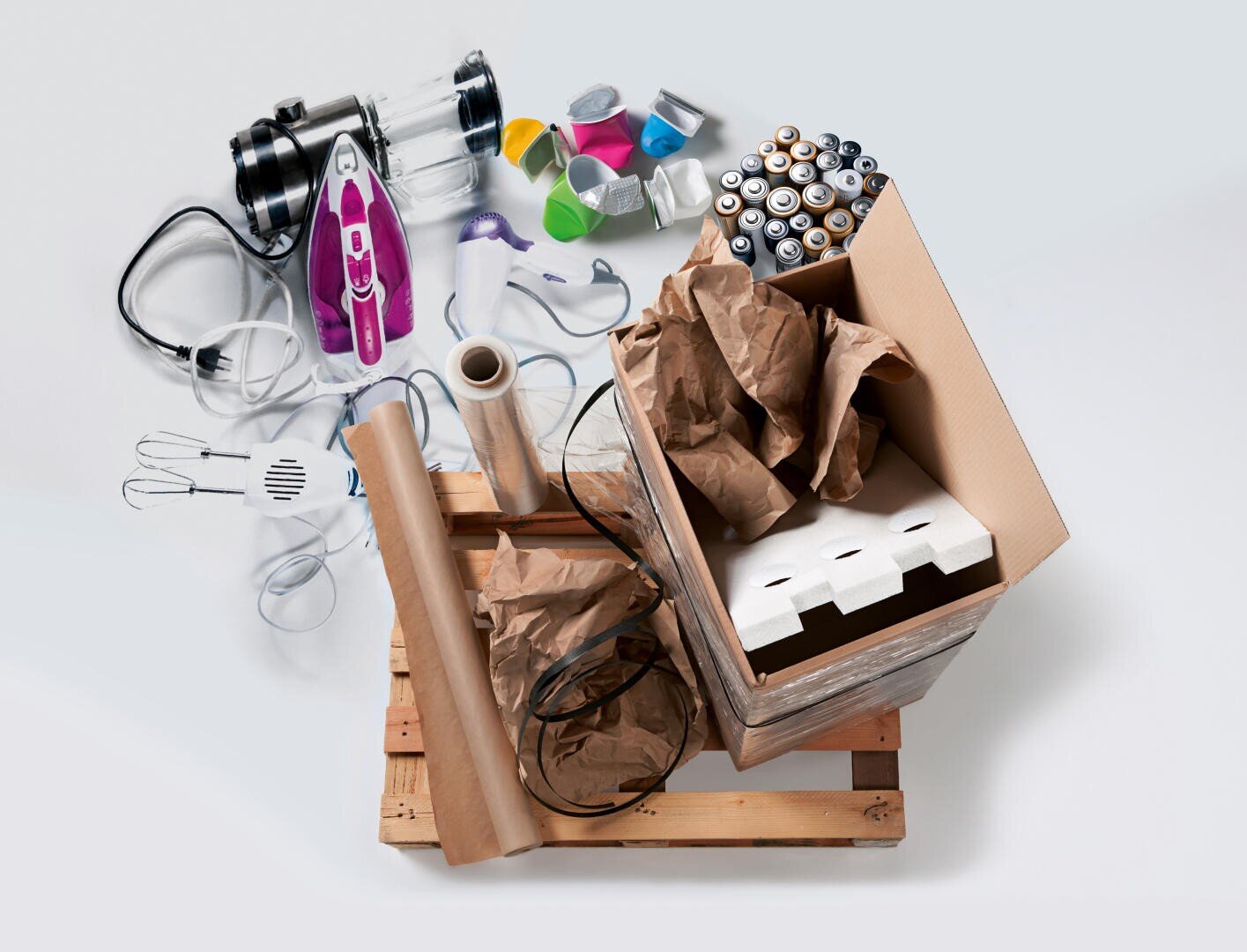 Auf einer Holzpalette steht eine Pappschachtel, die mit braunem Papier, Schaumstoff, Plastikfolie und Kabeln gefüllt ist. Daneben stehen Batterien, Kleingeräte, Farbdosen und ein lila Bügeleisen auf hellgrauem Hintergrund.