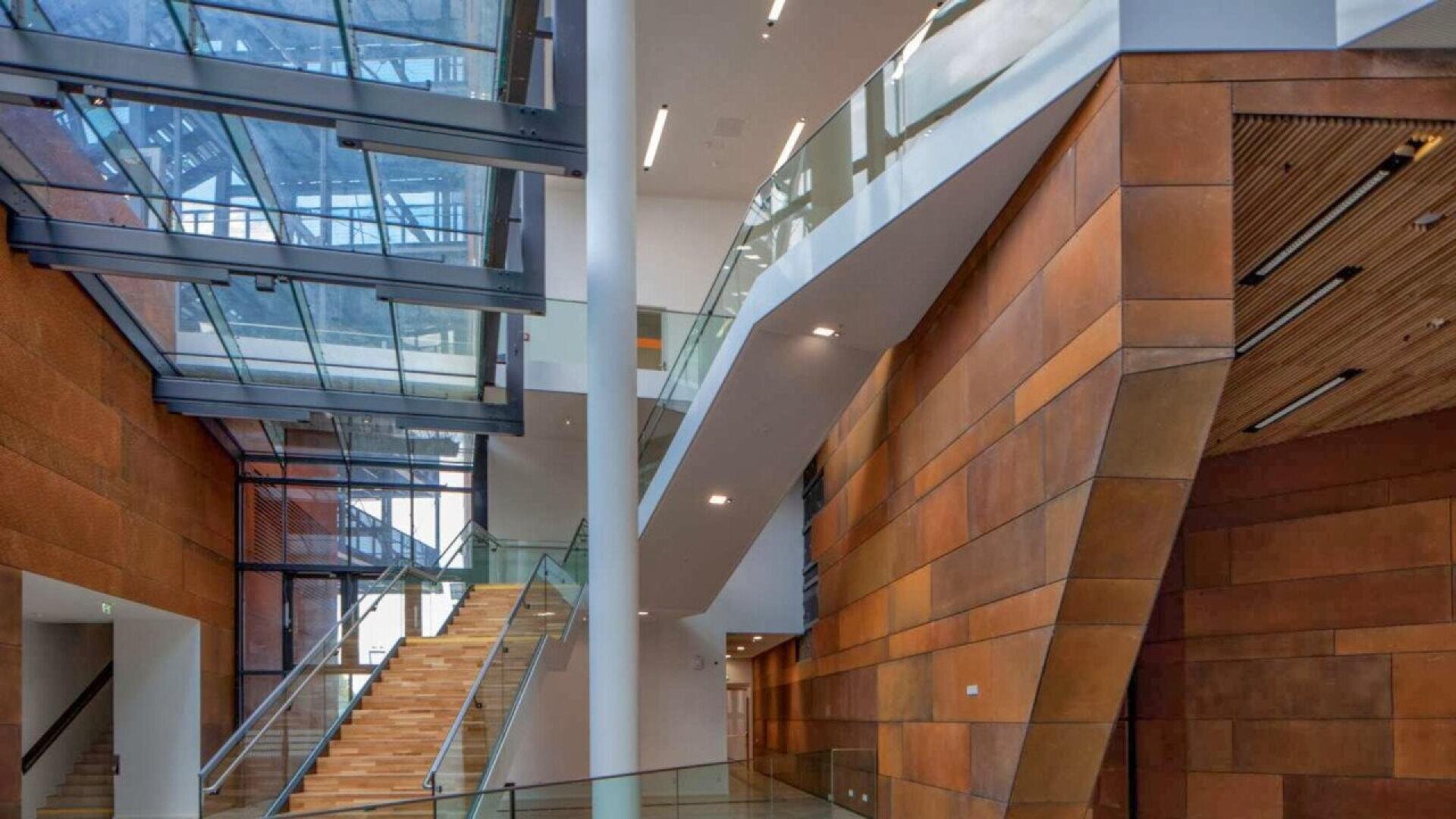Modern building interior with glass walls, a wide staircase with tan steps, brown panel walls, glass railings, and an angular ceiling structure letting in natural light.