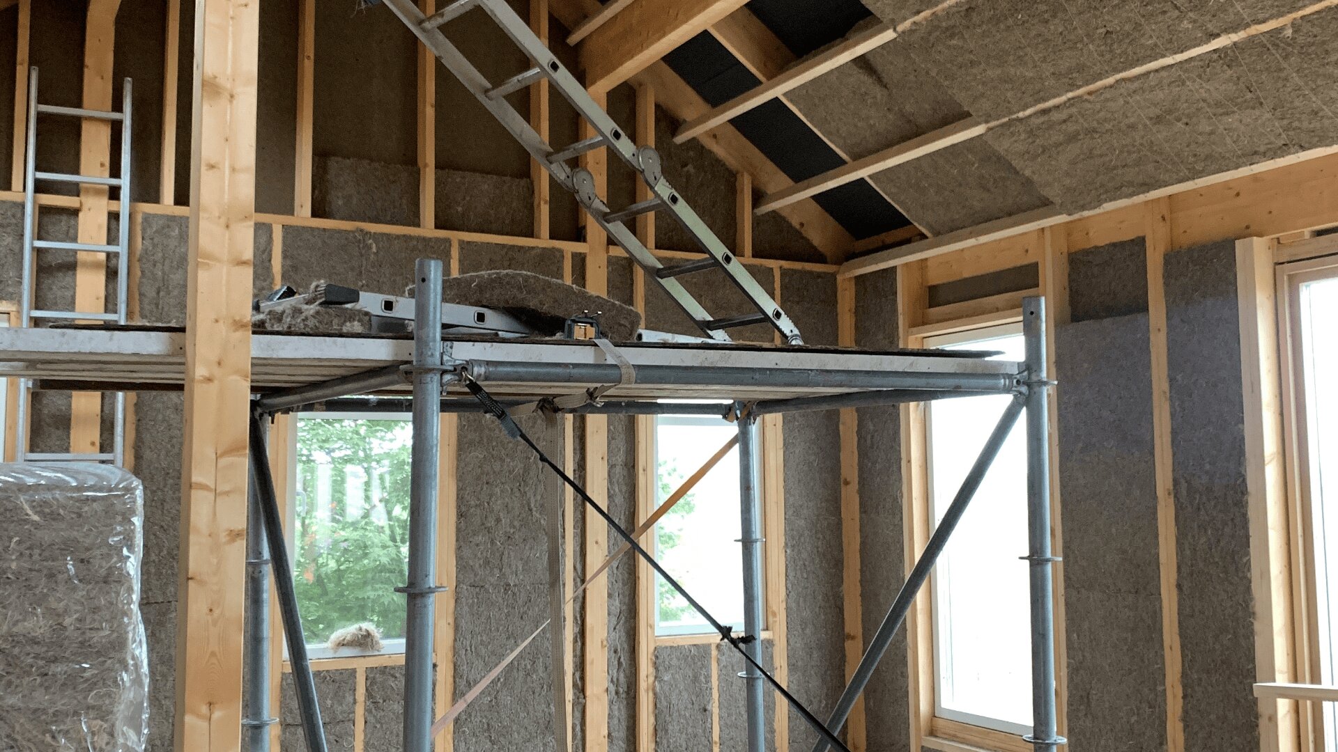 Im Bau befindliches Holzhaus mit sichtbarem Fachwerk, Dämmplatten in Wänden und Decke, Metallgerüst im Inneren und einer Leiter, die in einen unfertigen oberen Bereich führt. Natürliches Licht fällt durch große Fenster ein.