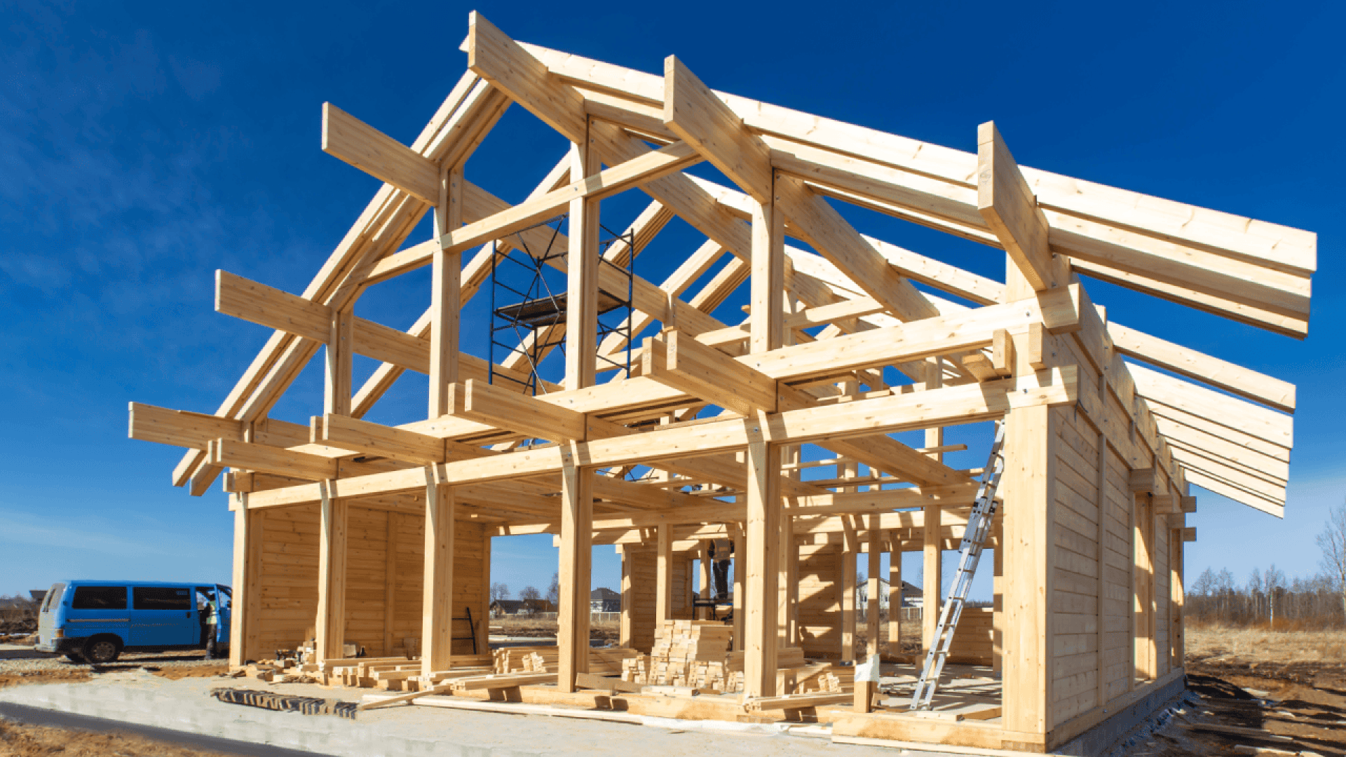 A wooden house frame under construction stands on a concrete foundation, with beams and supports visible. A ladder leans against the frame, and a blue van is parked nearby under a clear blue sky.